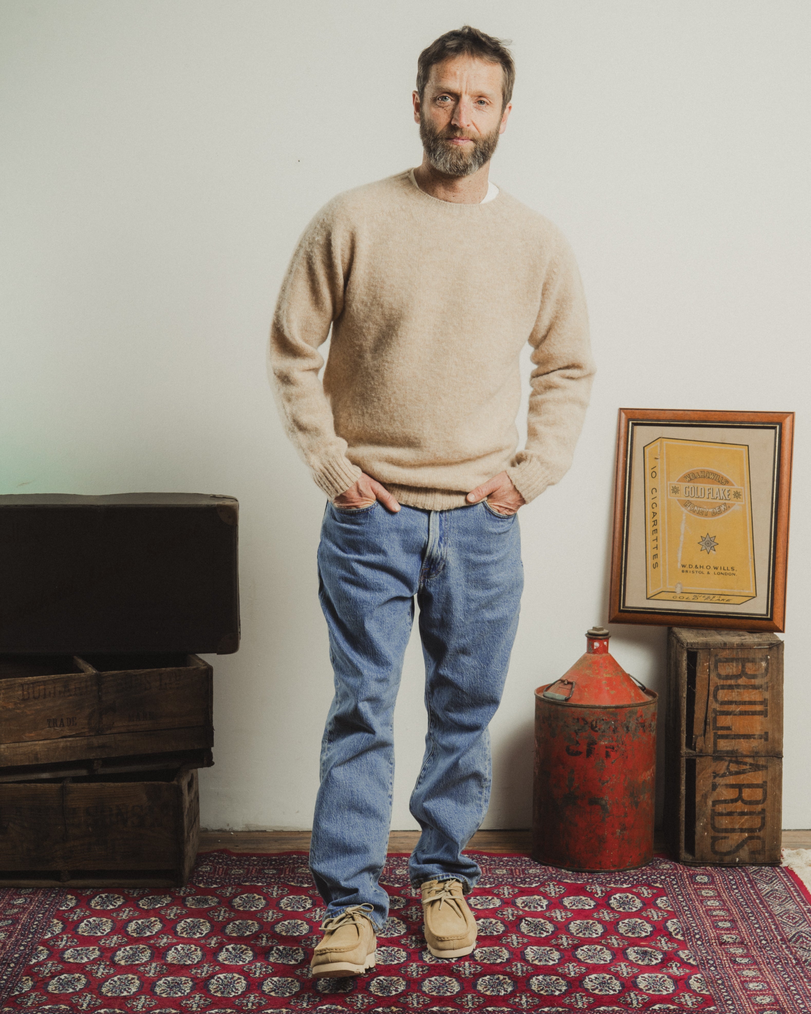Man wearing a beige sweater and blue jeans standing in a room with a red rug and wooden crates.