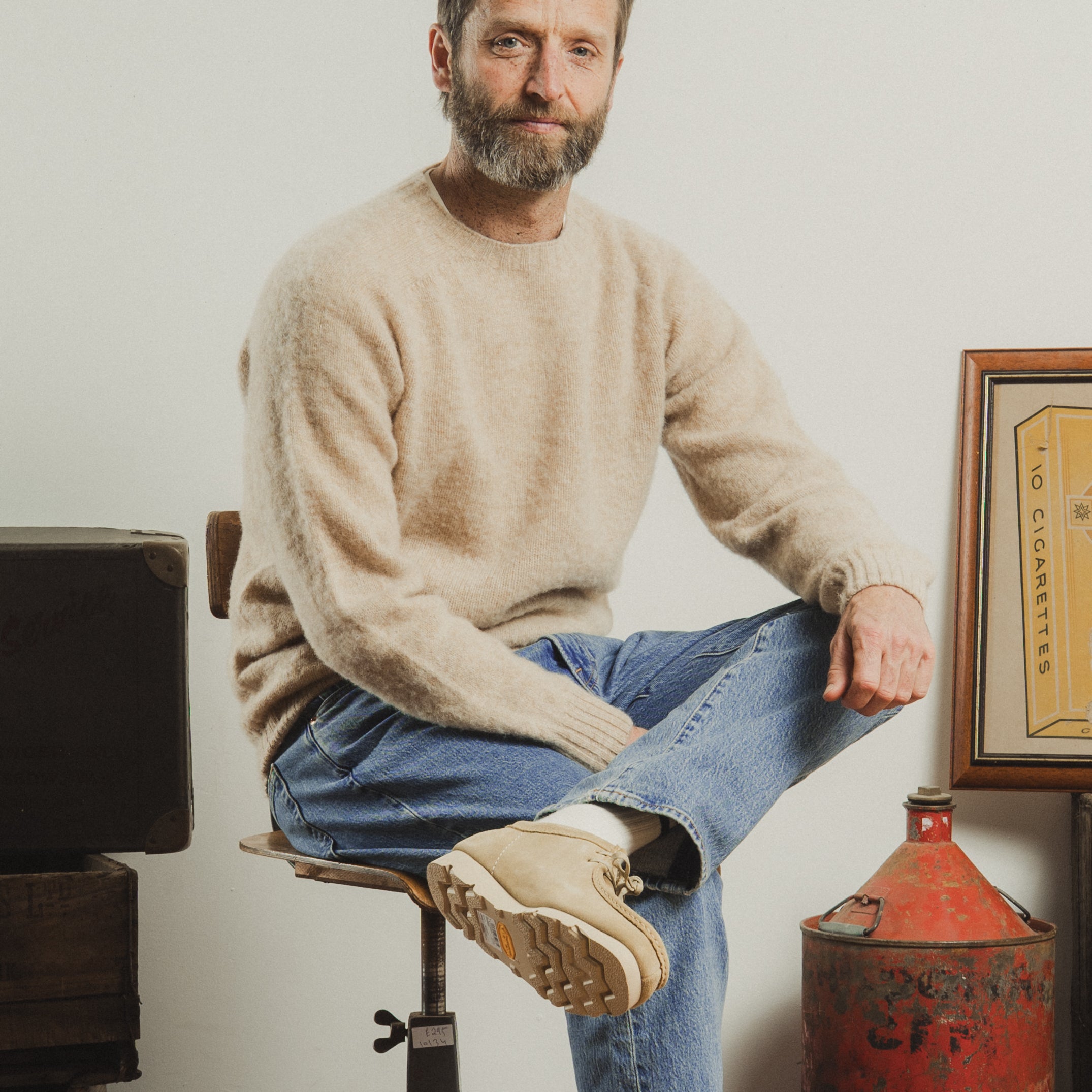 Man sitting on a stool wearing a beige sweater and blue jeans, with a plain background.