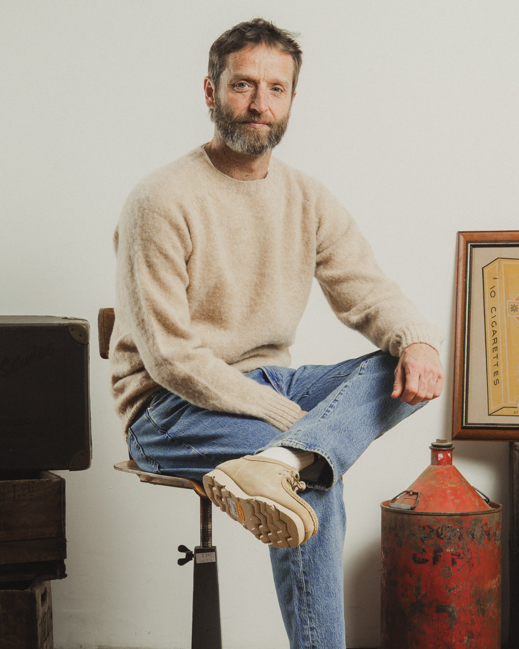 Man sitting on a stool wearing a beige sweater and blue jeans, with a plain background.