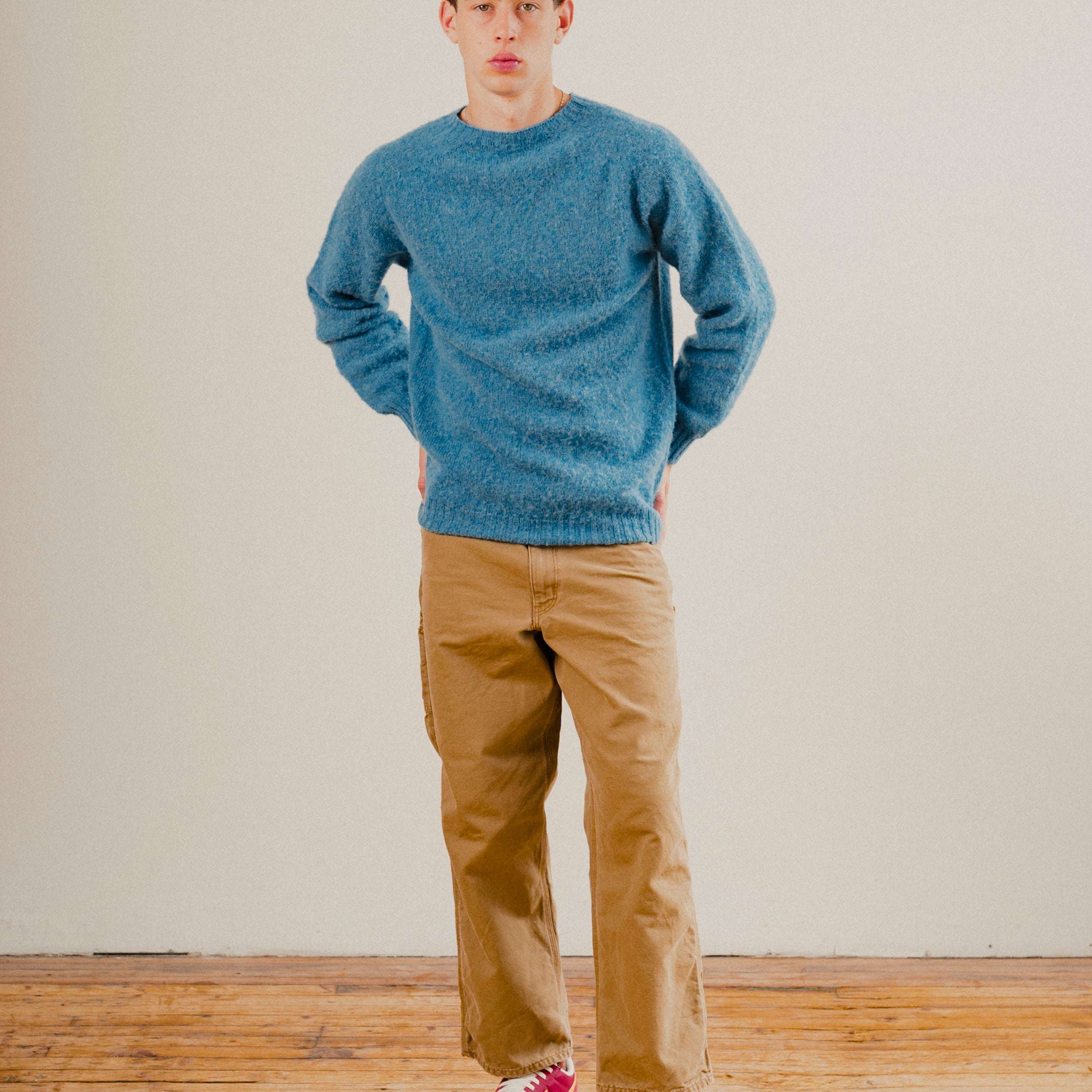 Man in a blue wool knit crewneck jumper sweatshirt on a plain background with a wooden floor