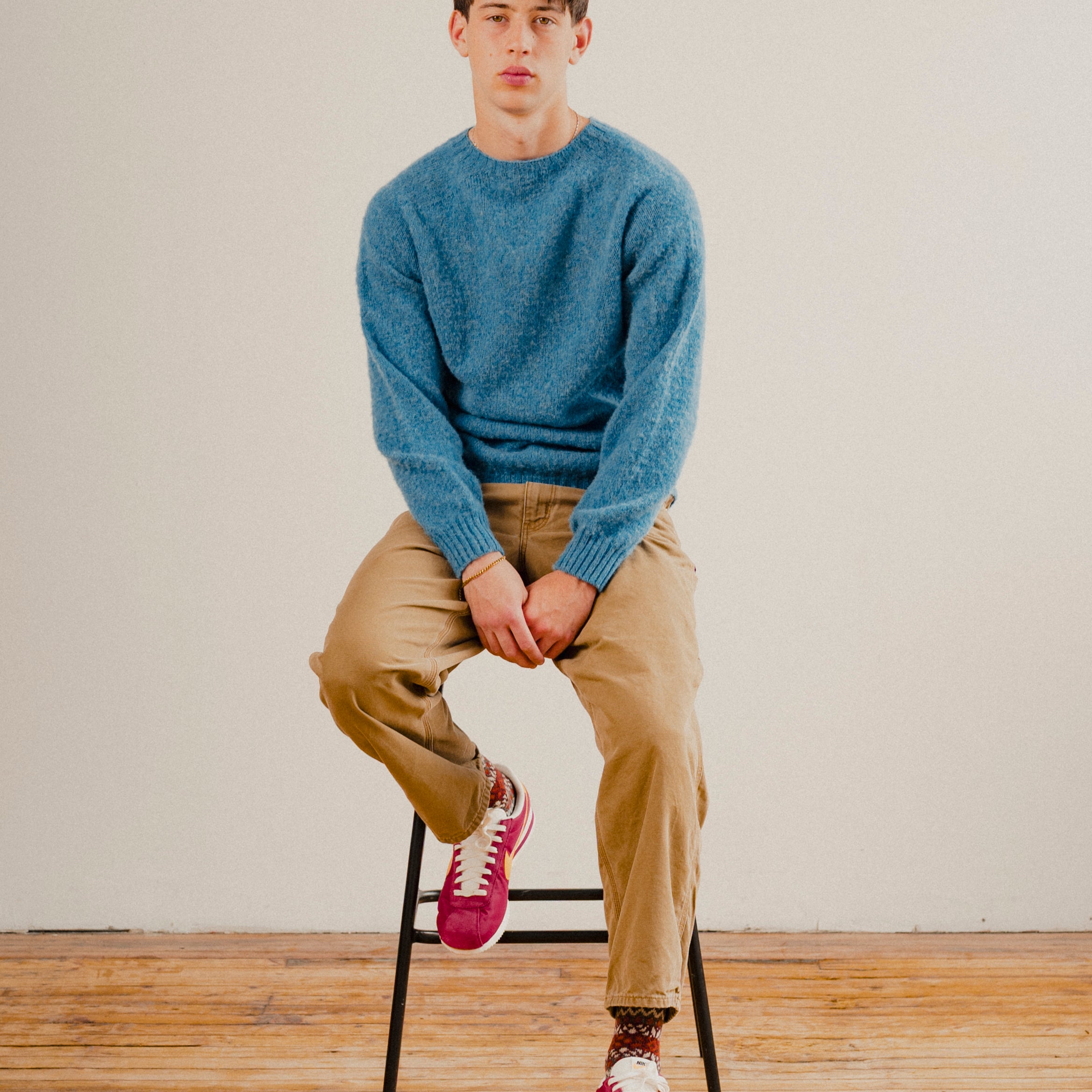 Man sat on stool in a blue wool knit crewneck jumper sweatshirt with a wooden floor and a plain white background
