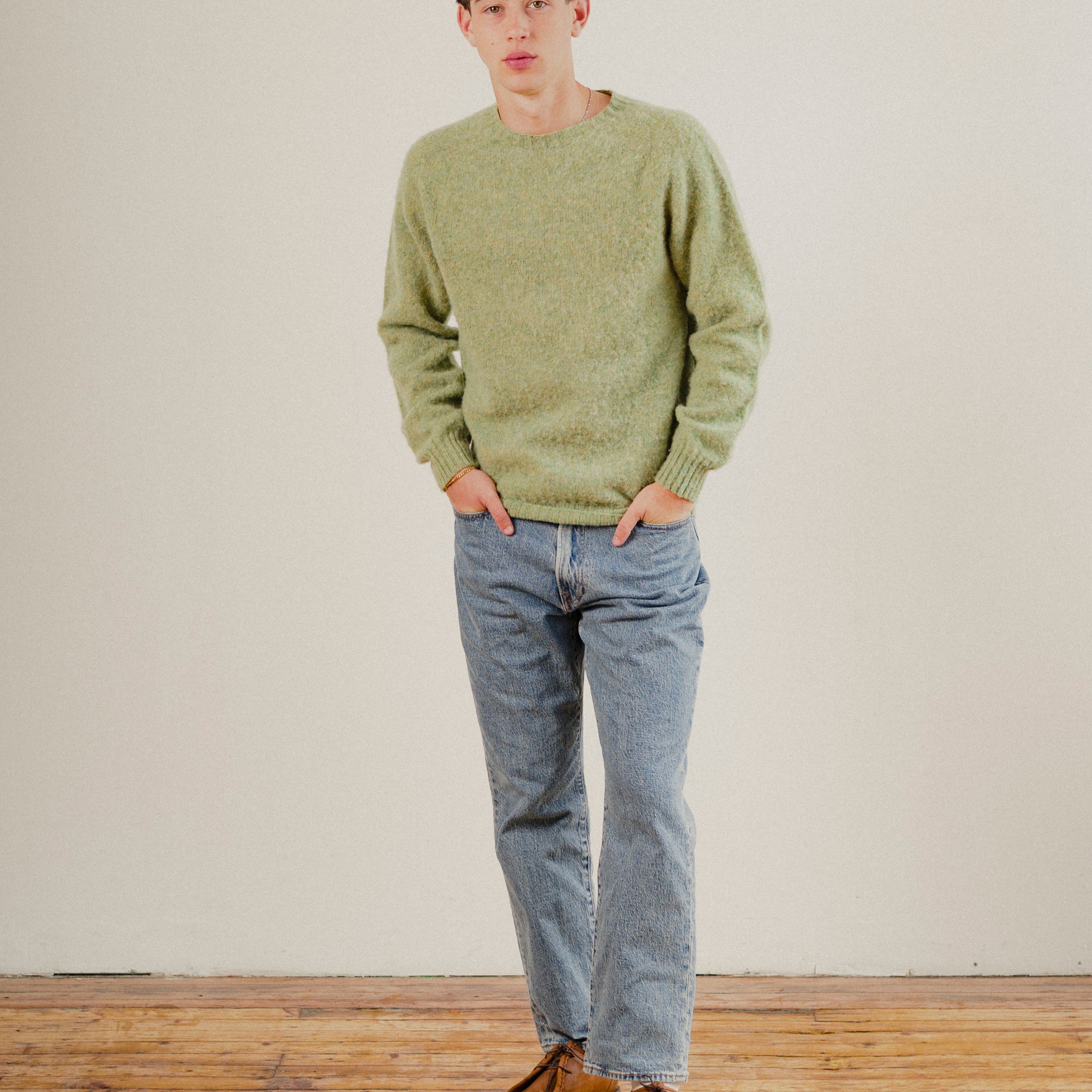 Man wearing a green wool knit sweater and blue jeans standing on a wooden floor with a white wall background