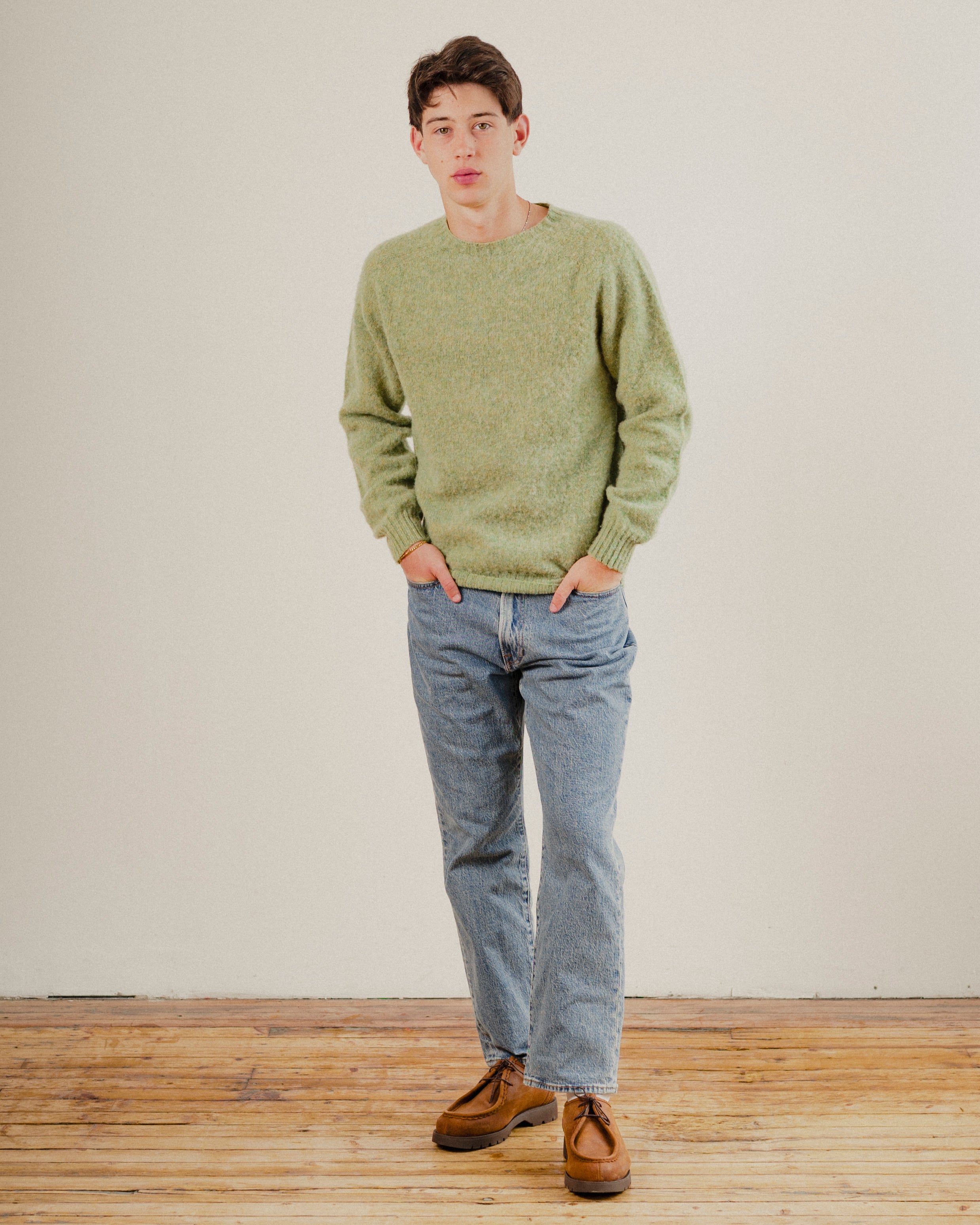Man wearing a green wool knit sweater and blue jeans standing on a wooden floor with a white wall background