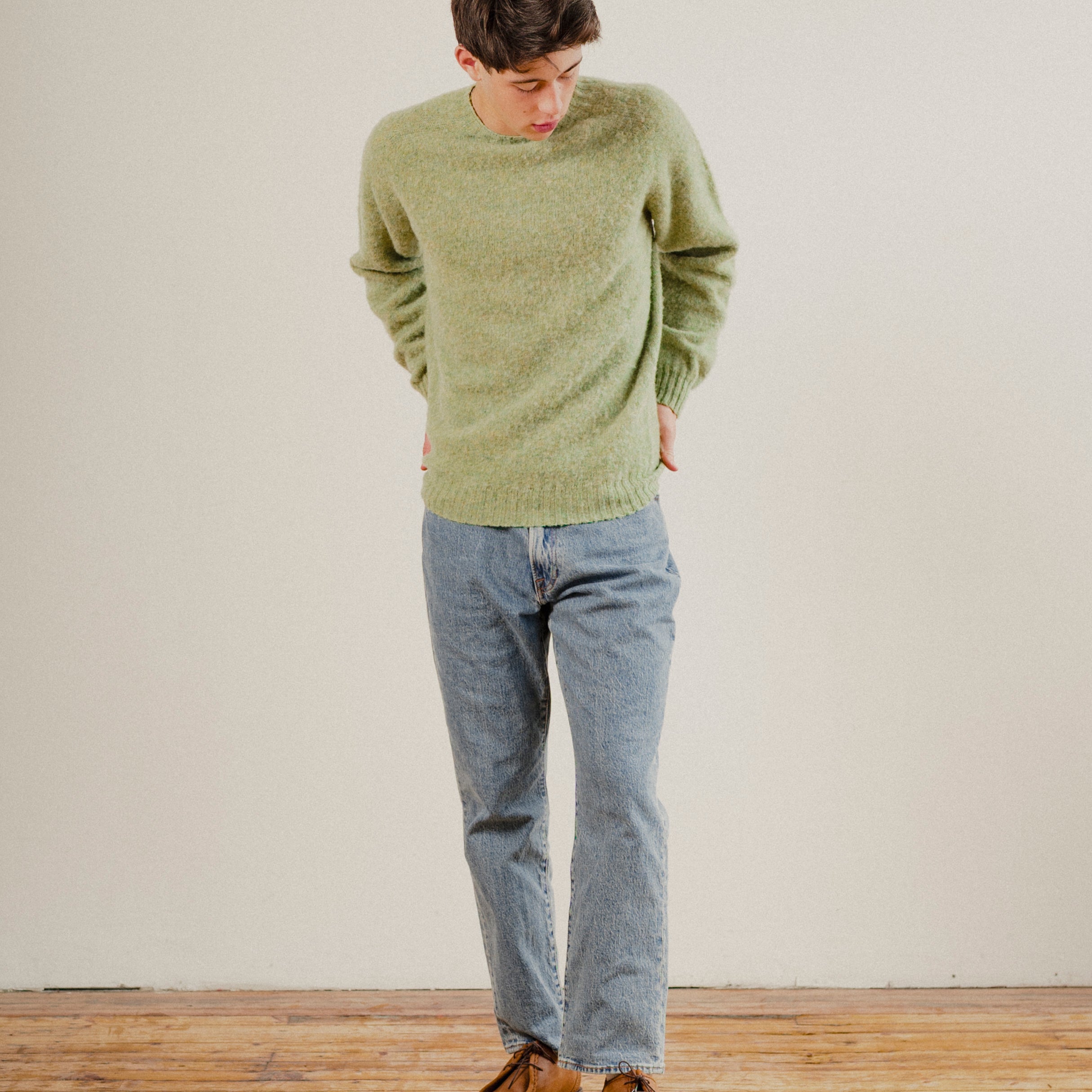 Person wearing a green wool knit sweater and blue jeans standing on a wooden floor with a white wall background