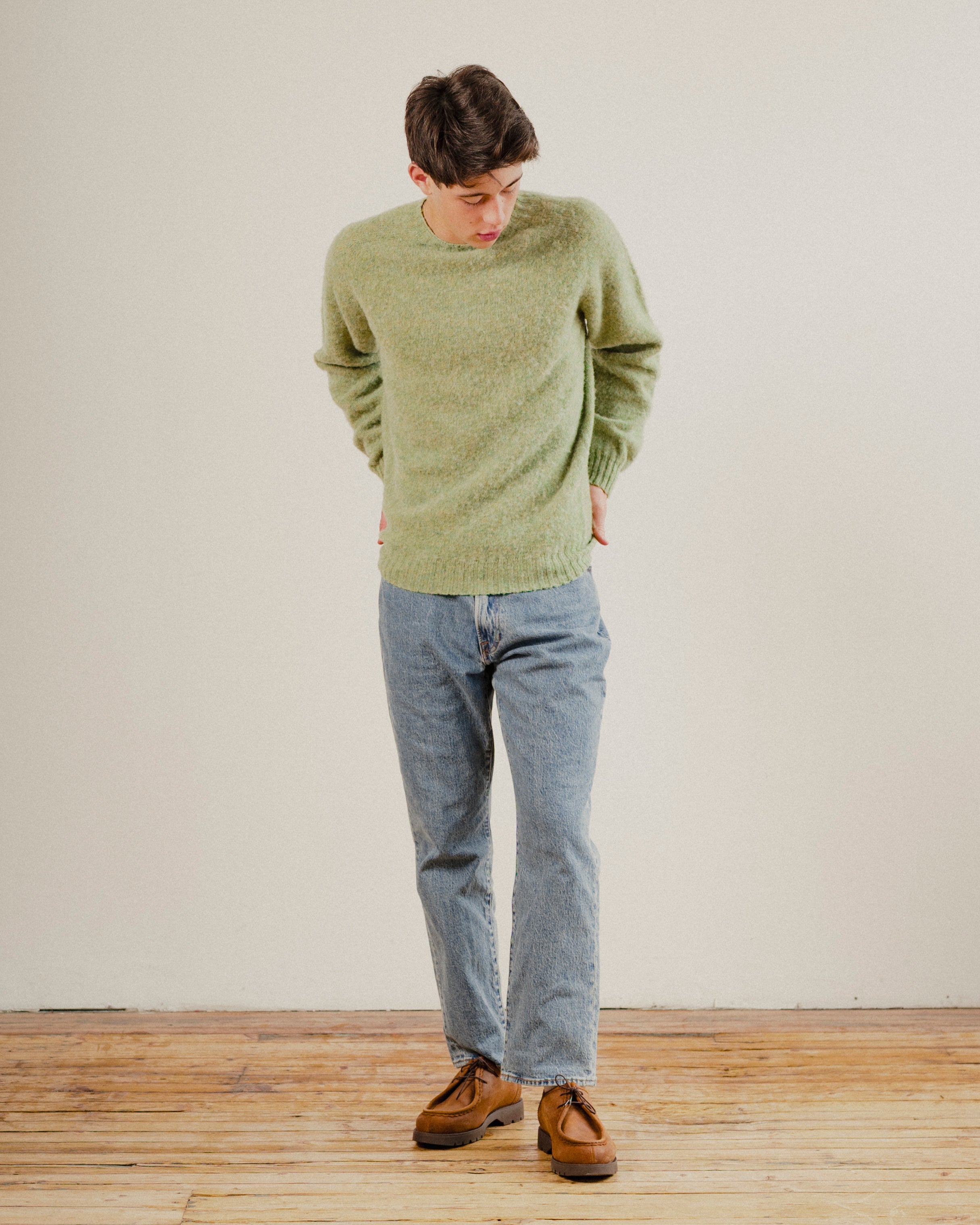 Person wearing a green wool knit sweater and blue jeans standing on a wooden floor with a white wall background