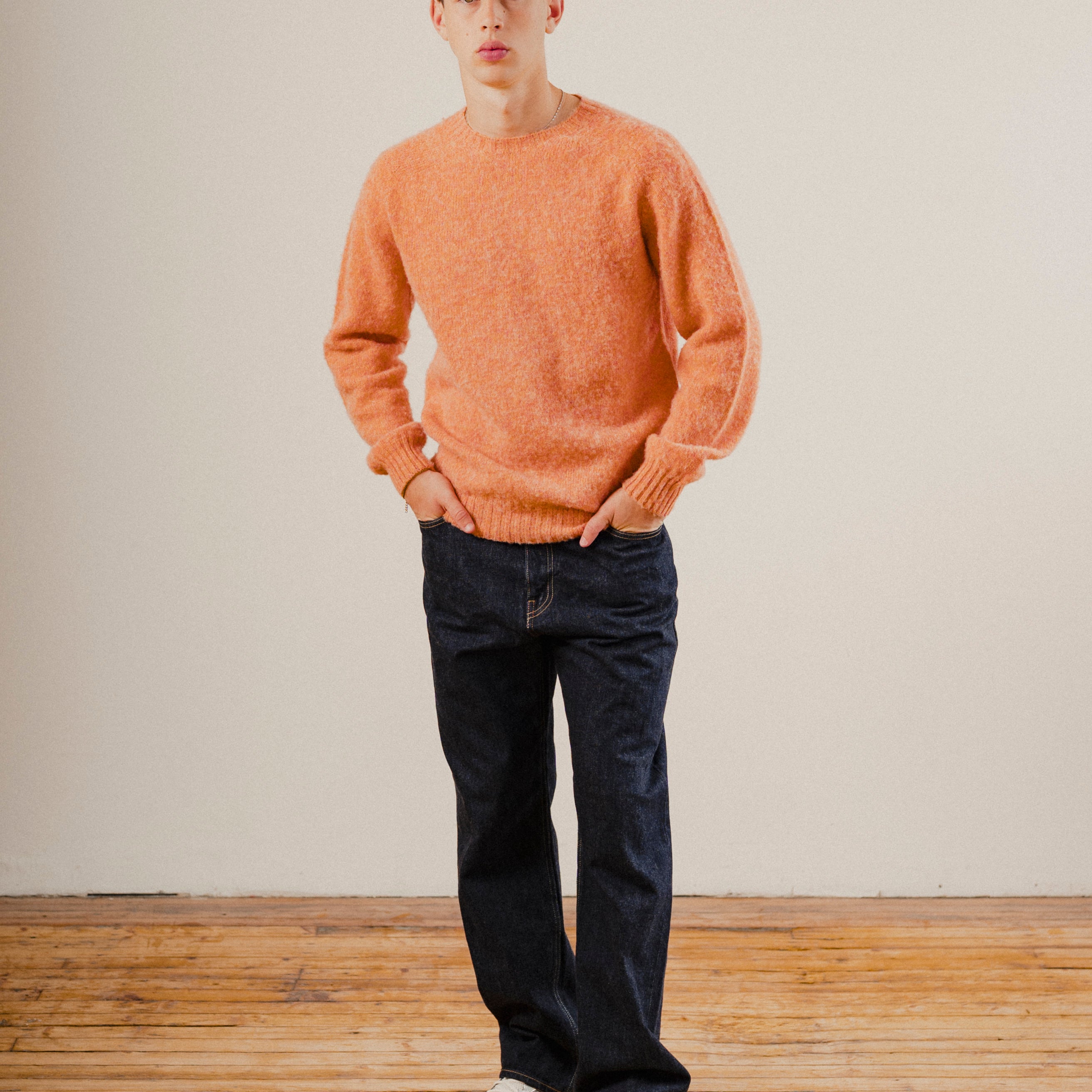Man wearing orange wool knit sweater with dark blue denim jeans on a wooden floor with a white background