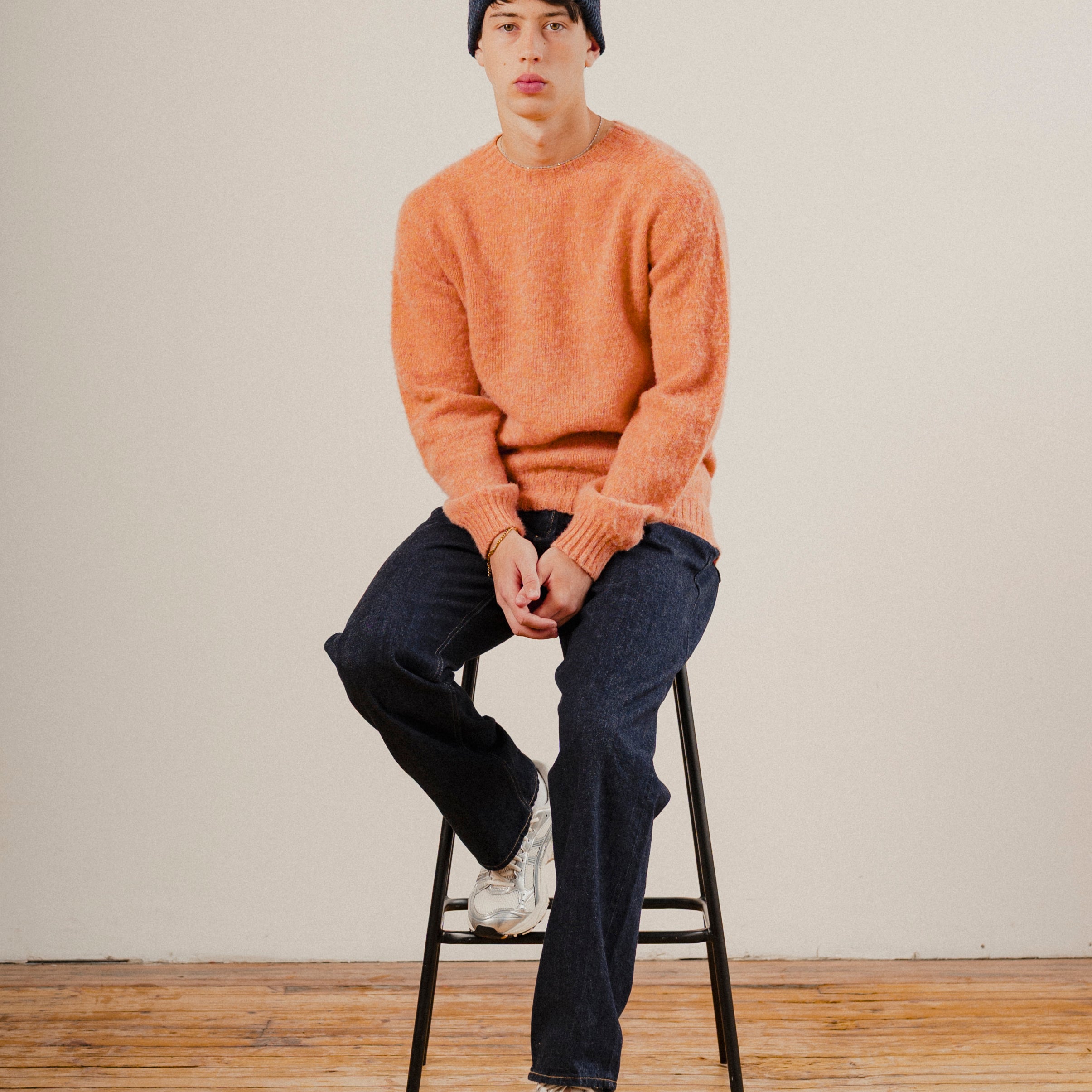 man sat on a stool wearing an orange wool knit crewneck with dark blue denim jeans and a navy blue wool beanie