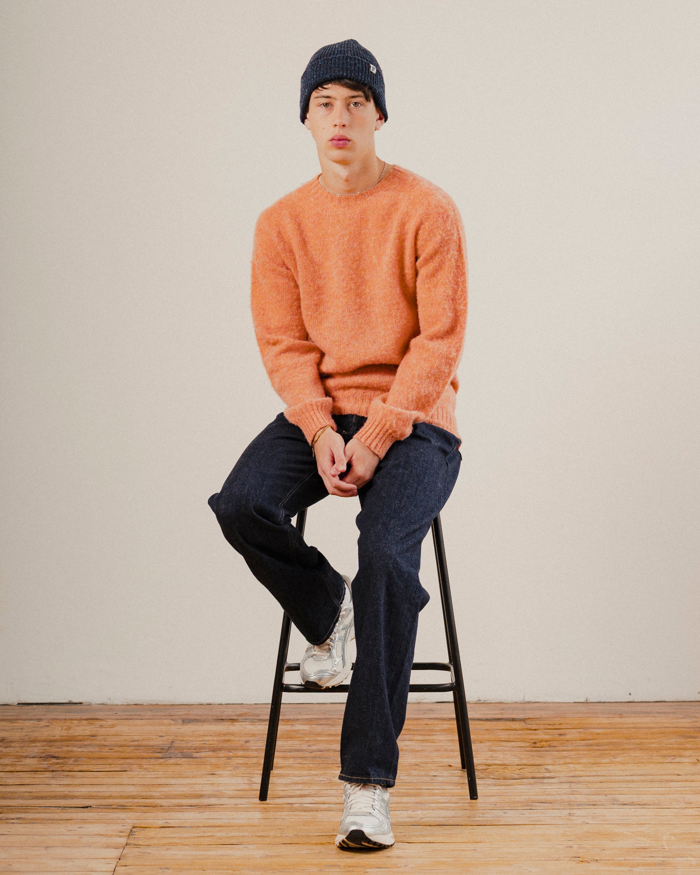 man sat on a stool wearing an orange wool knit crewneck with dark blue denim jeans and a navy blue wool beanie