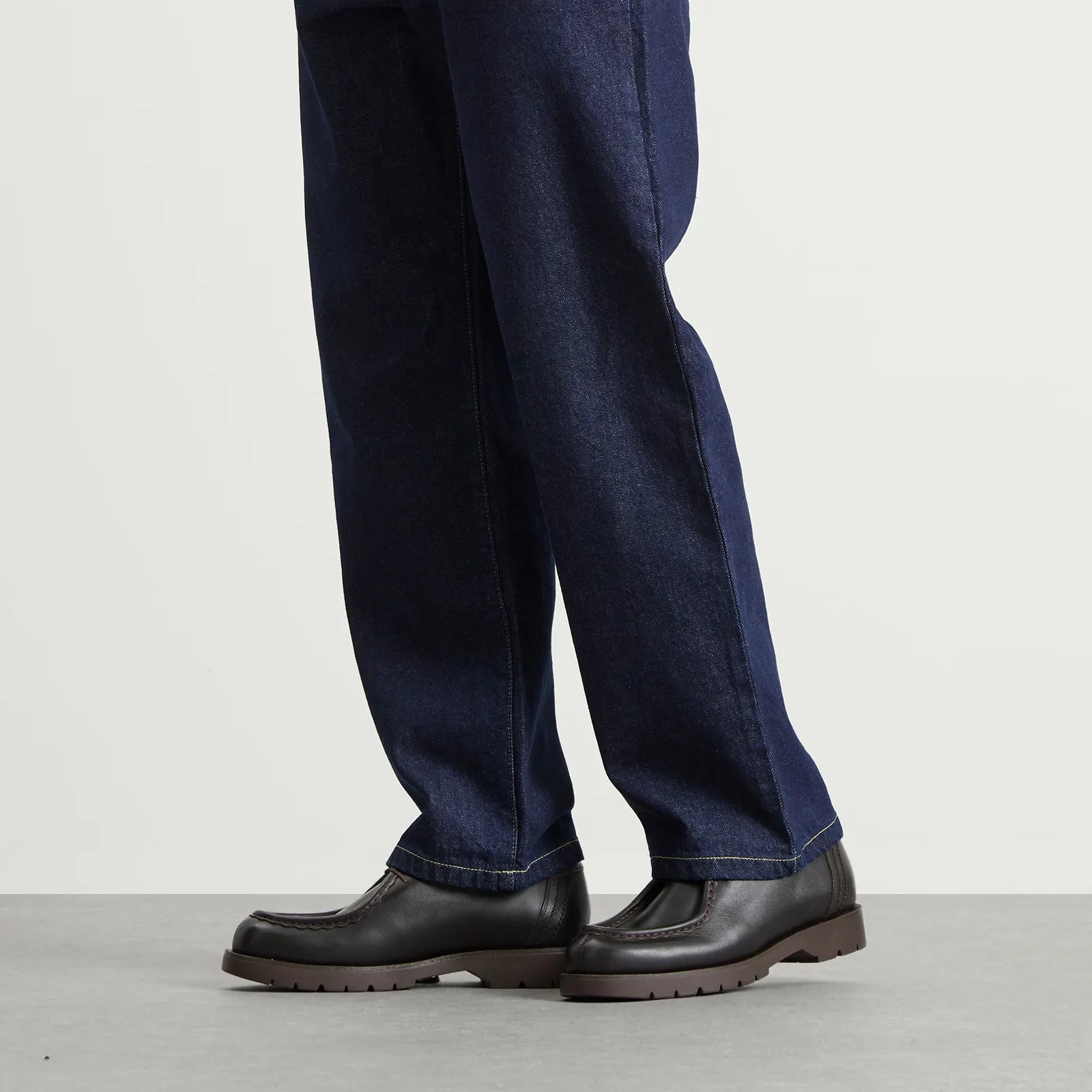 Person wearing dark blue jeans and brown Kleman shoes on a light gray background