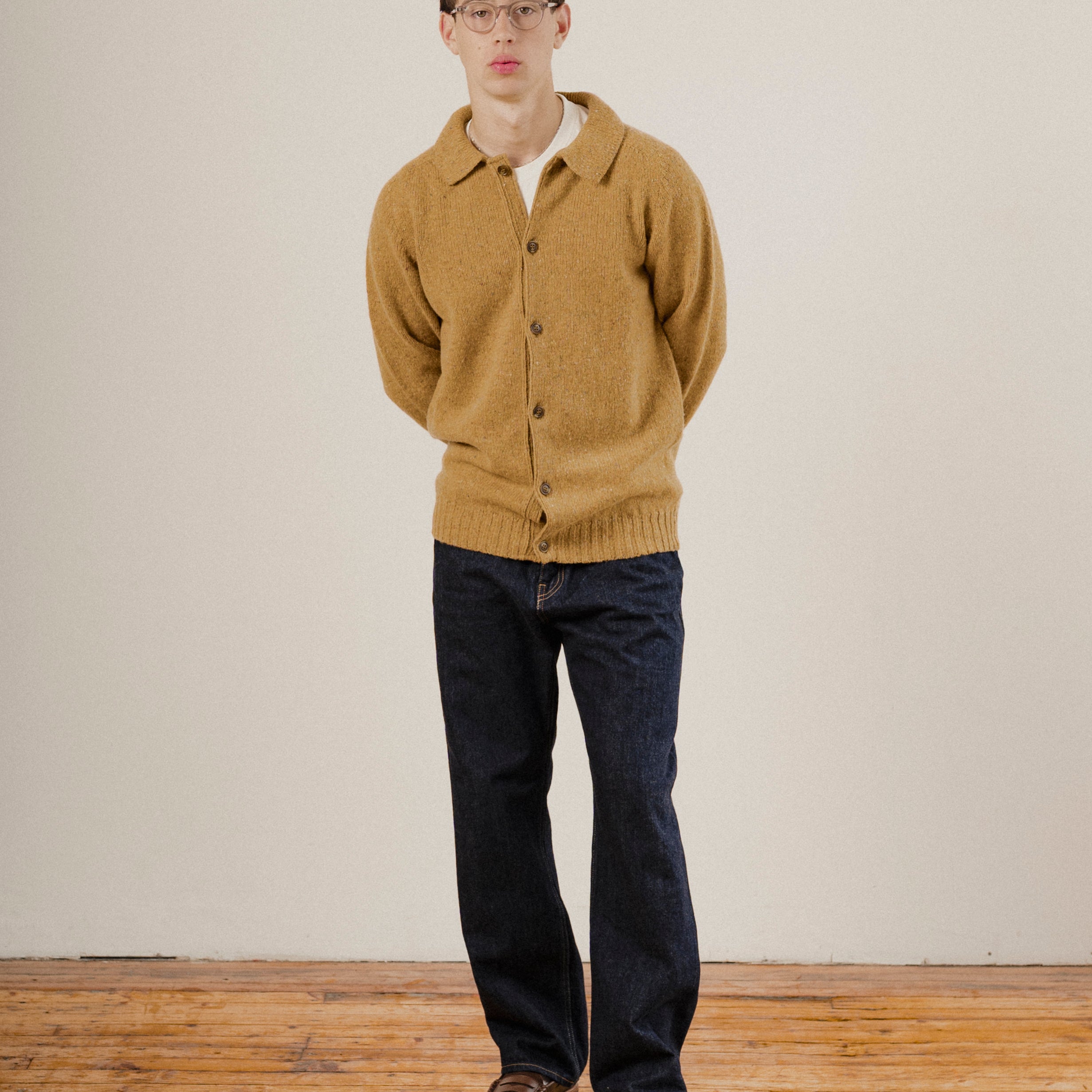 Man wearing a mustard yellow wool cardigan and dark jeans standing against a plain white wall.