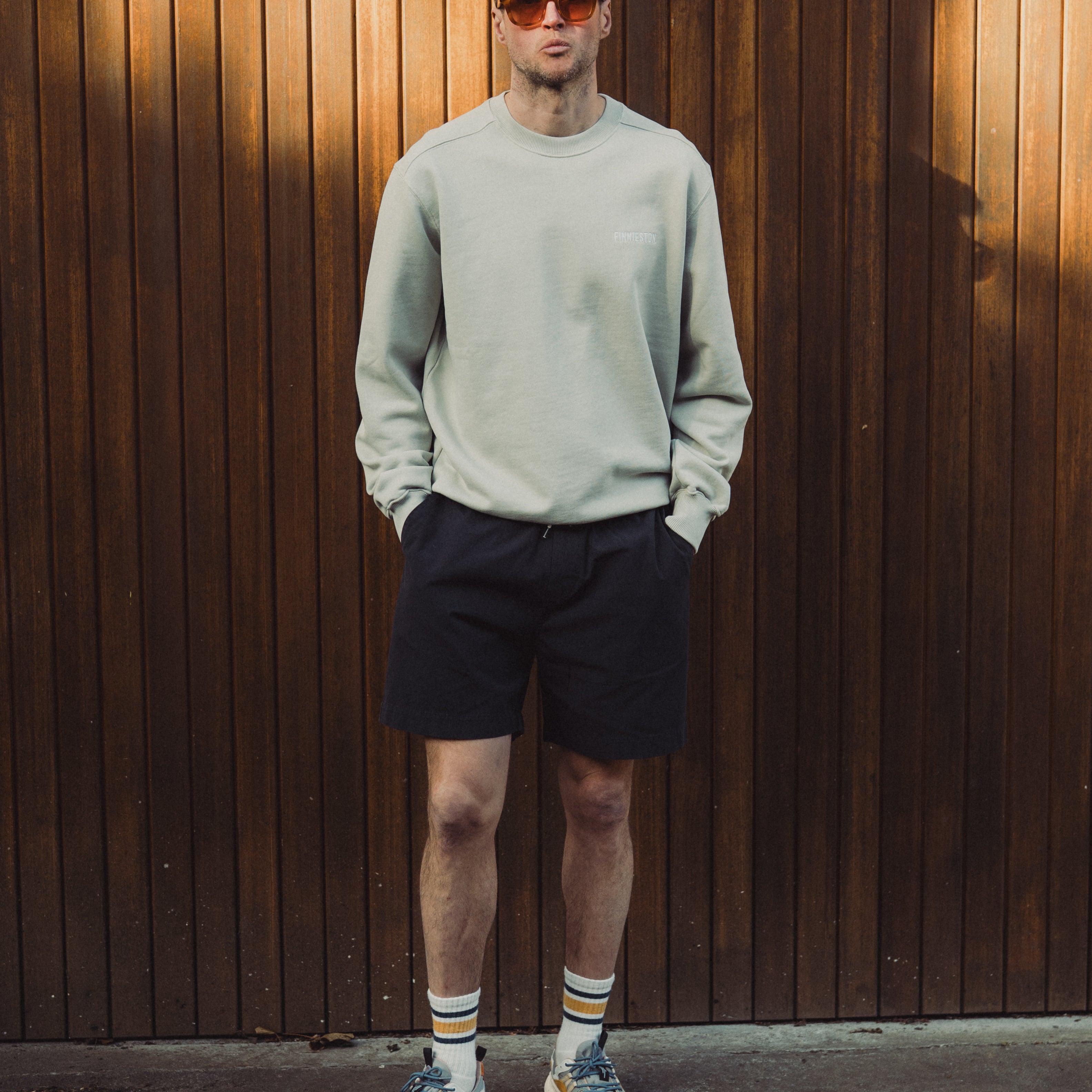 Man wearing a light gray sweatshirt, black shorts, and sunglasses standing against a wooden wall.
