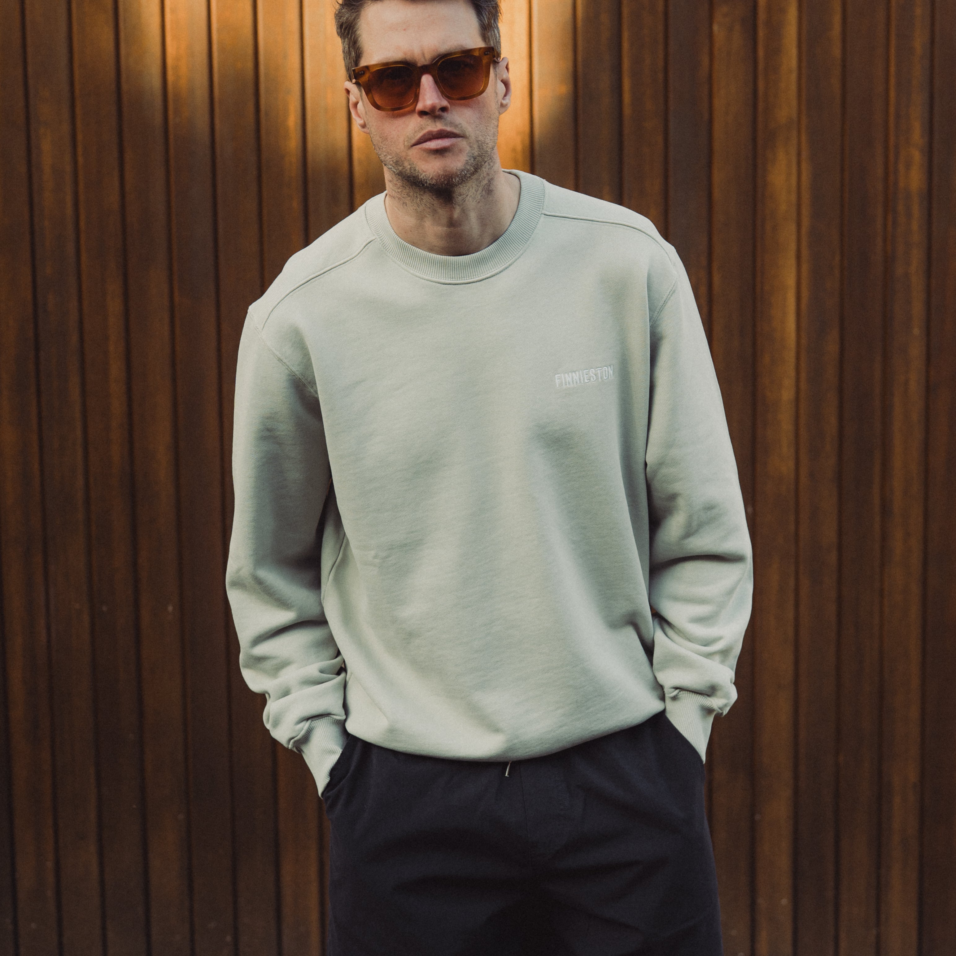 Man wearing a light gray sweatshirt and black shorts standing against a wooden wall.