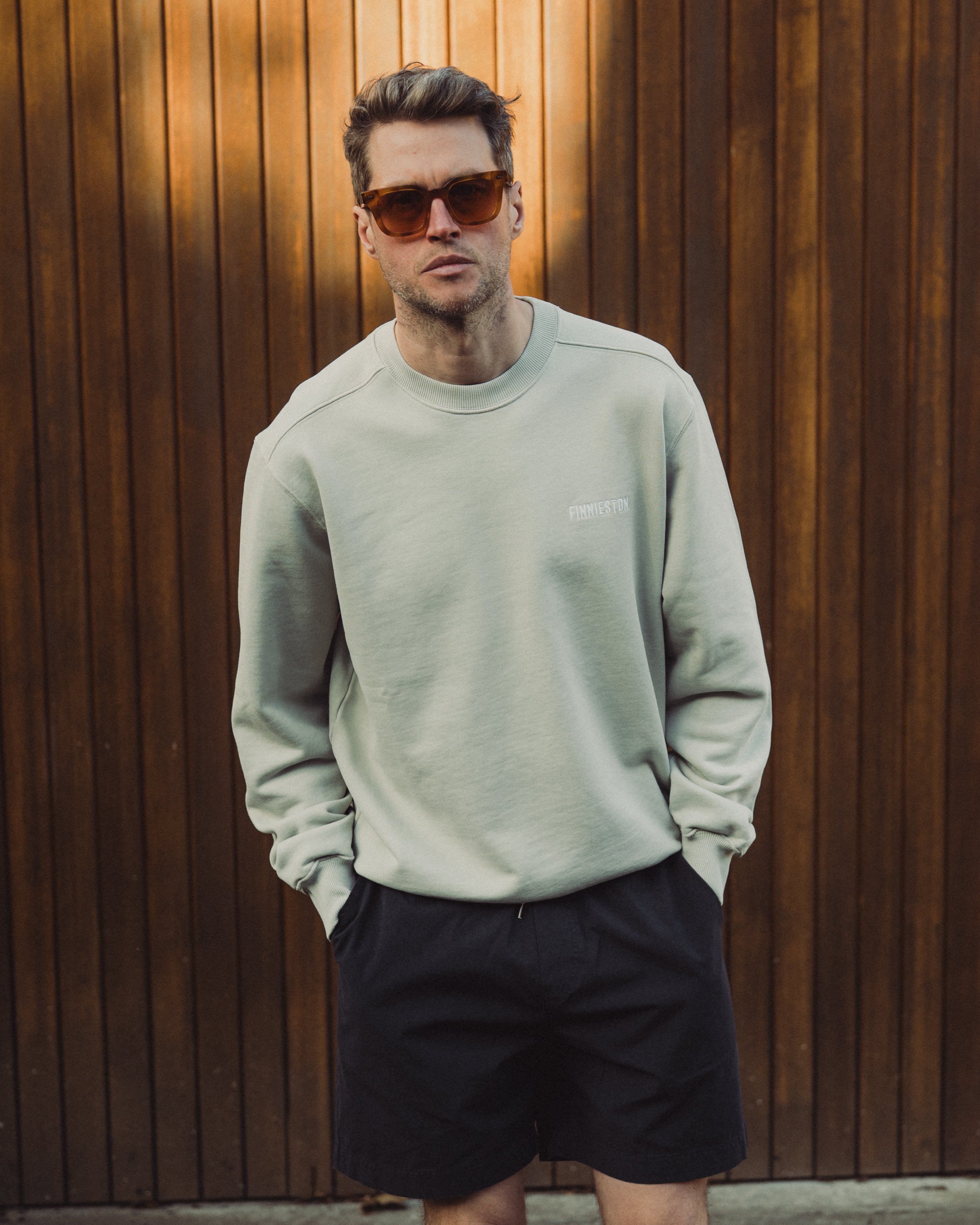 Man wearing a light gray sweatshirt and black shorts standing against a wooden wall.
