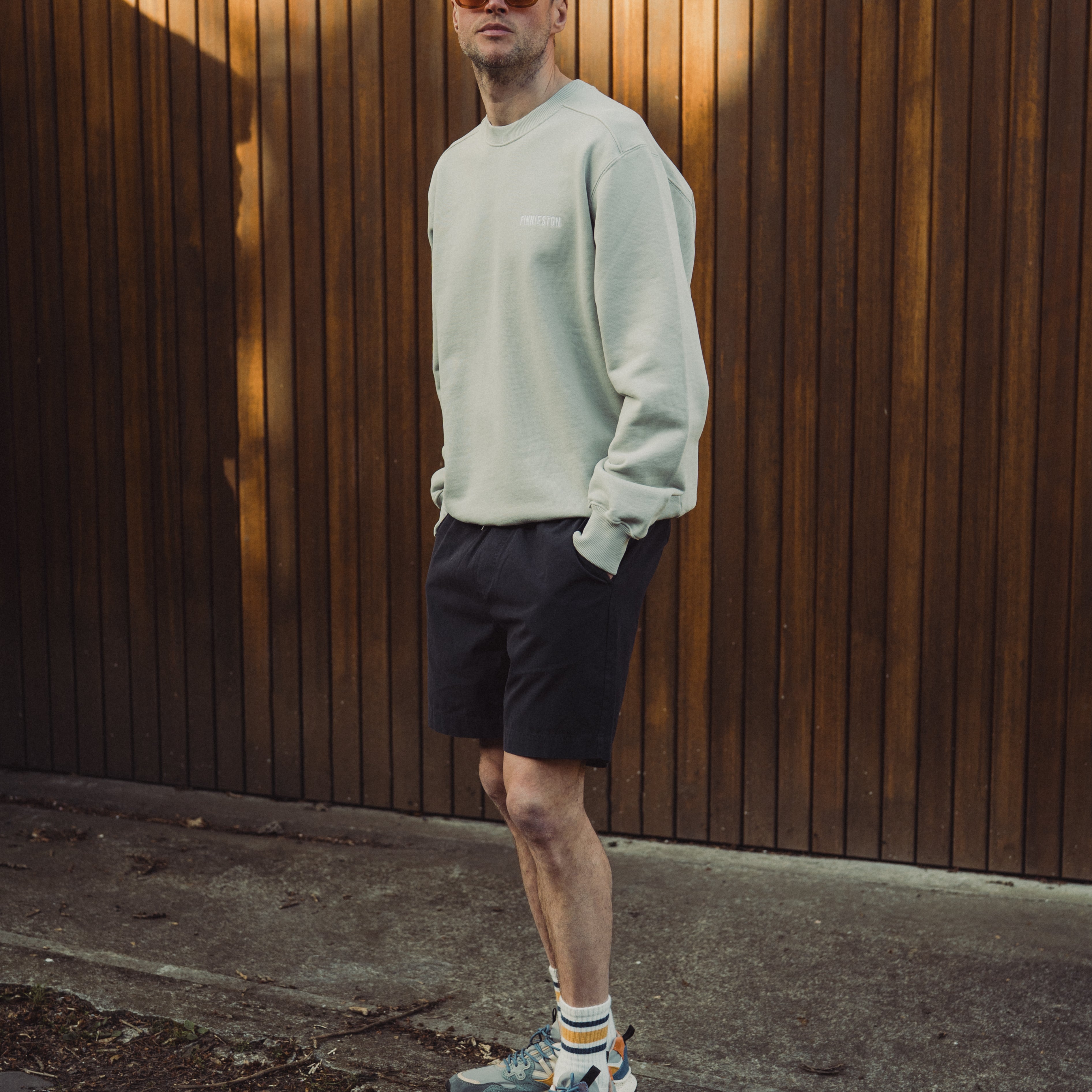 Man wearing a light gray sweatshirt and black shorts standing against a wooden wall.