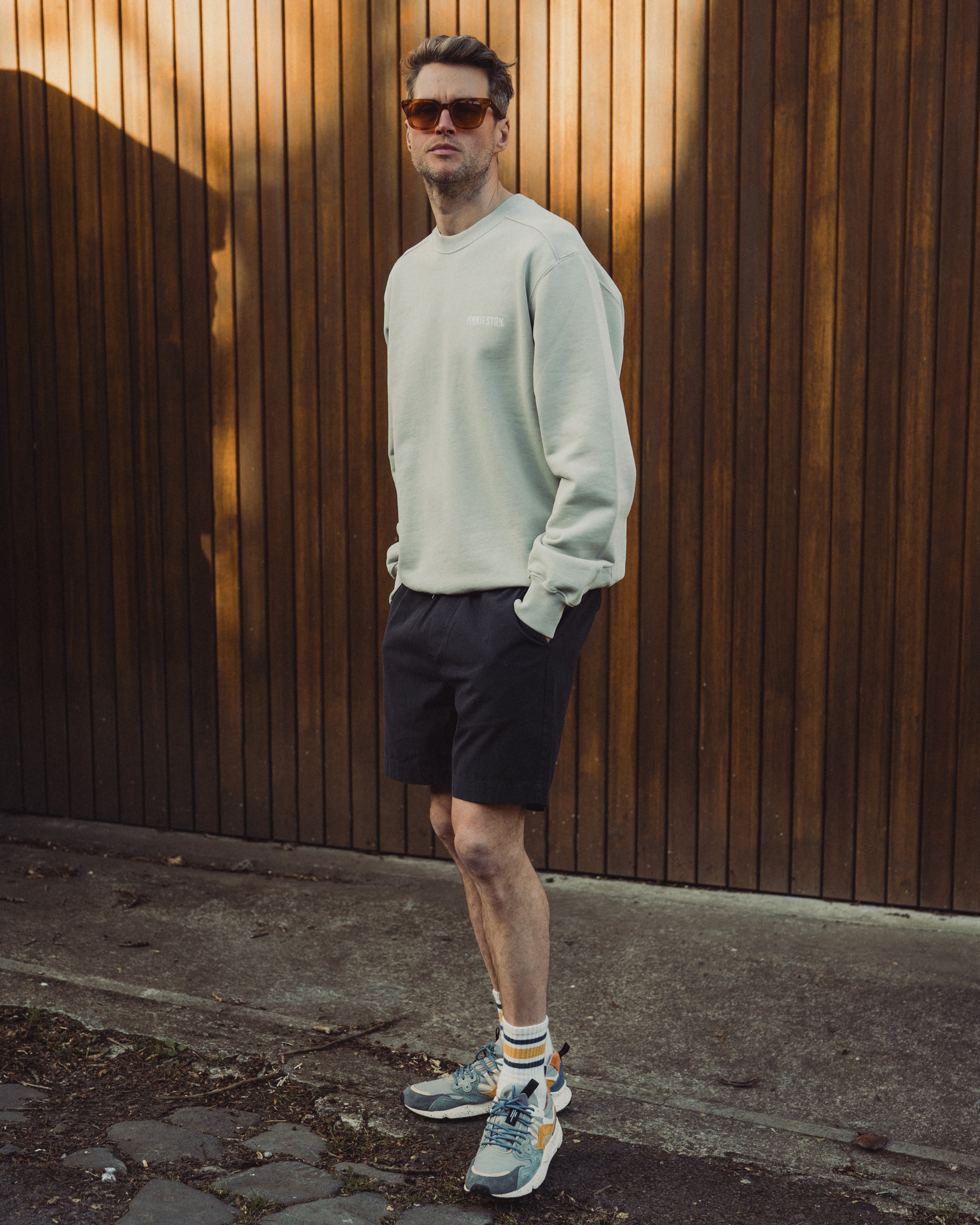 Man wearing a light gray sweatshirt and black shorts standing against a wooden wall.