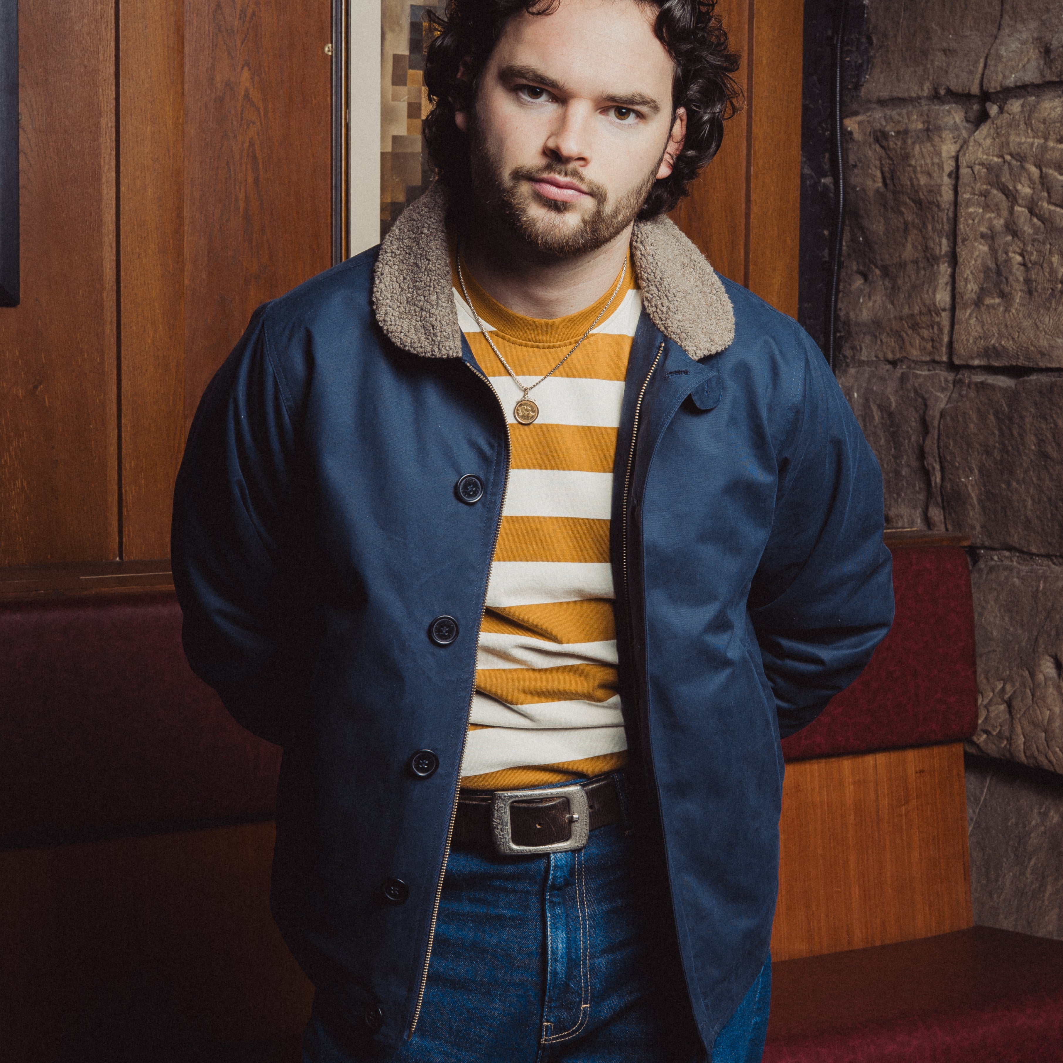 Man wearing a blue coat with a fur-lined hood, striped sweater, and jeans in an indoor setting.