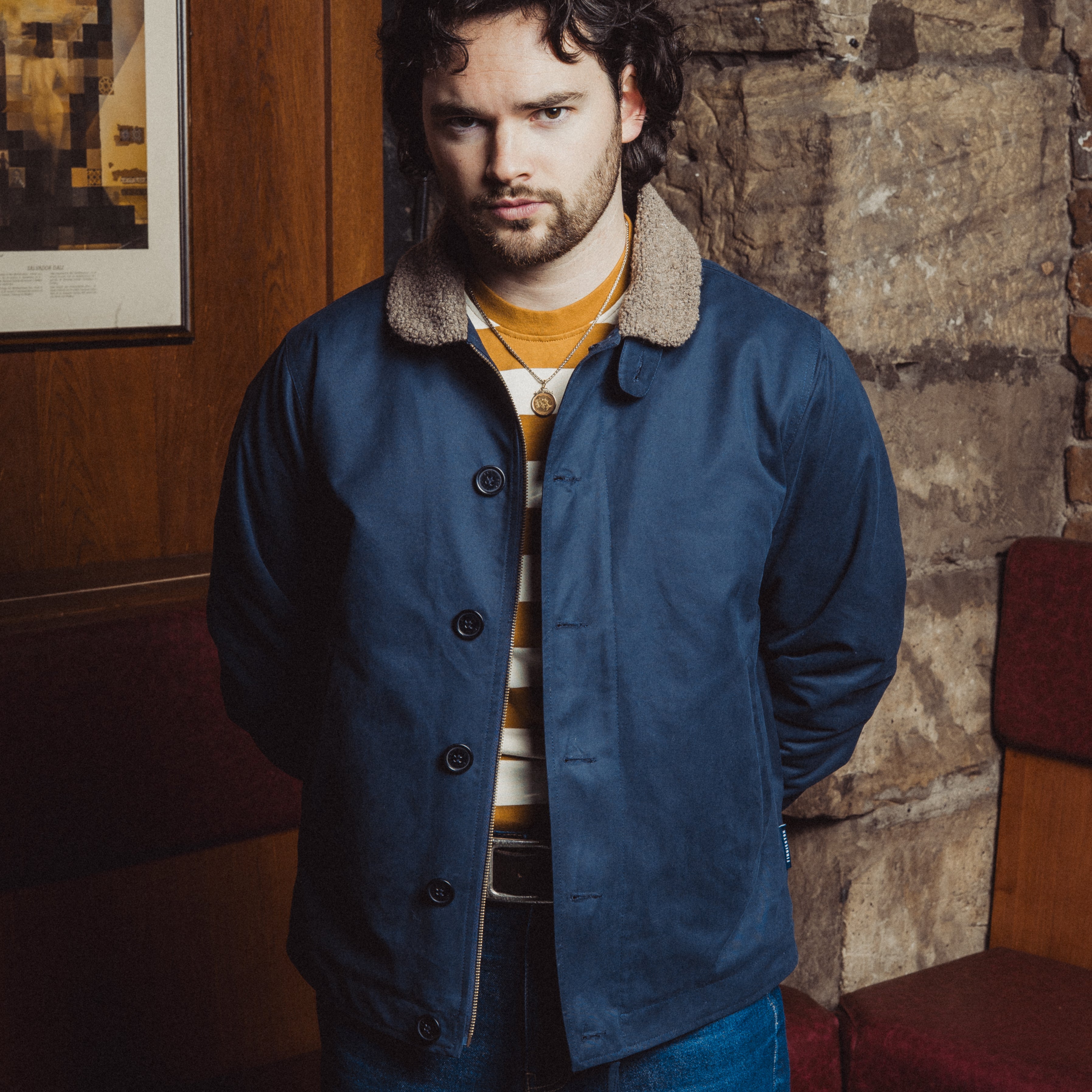 Man wearing a blue coat with a fur-lined hood in a room with stone walls and a framed picture.