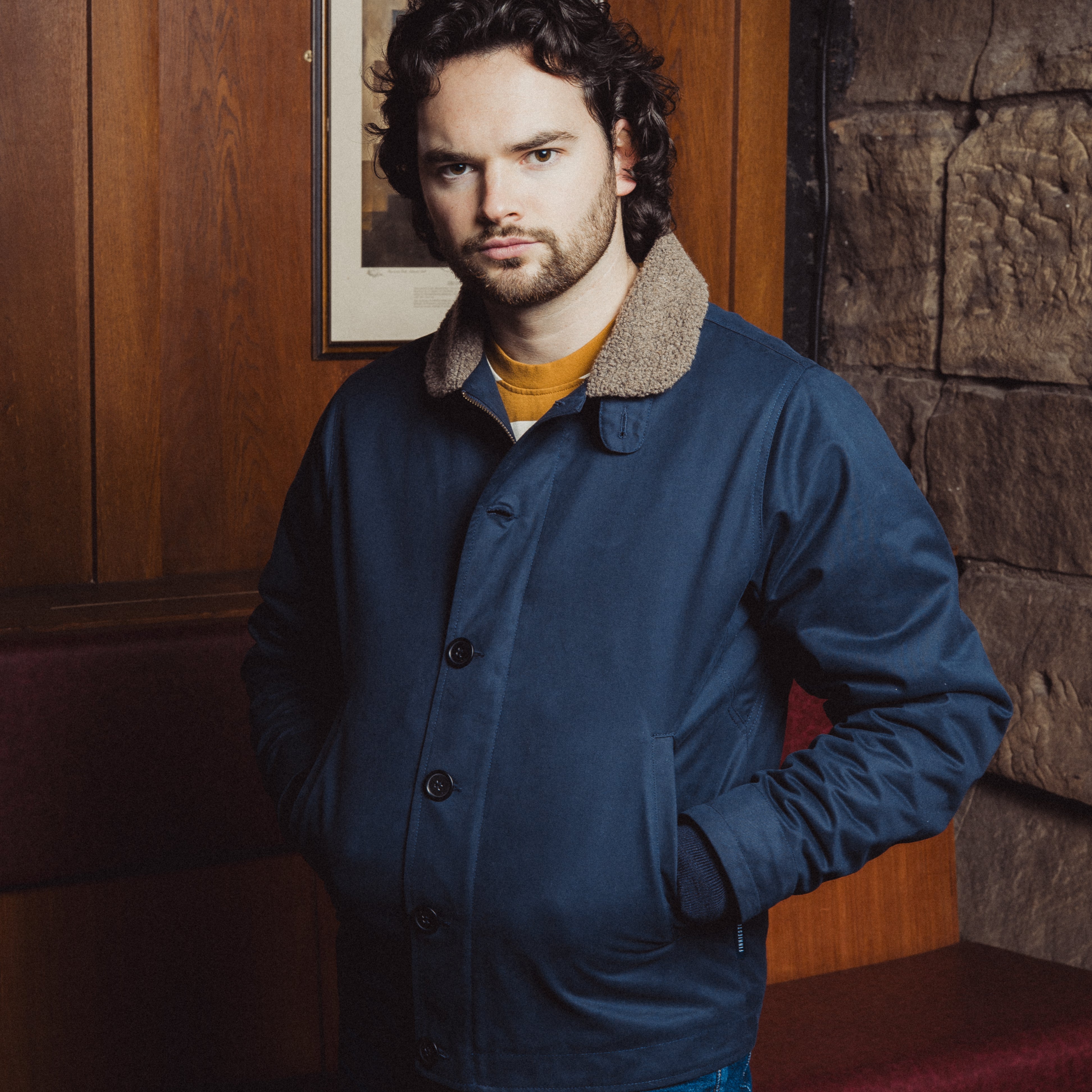 Man wearing a blue jacket in a room with wooden paneling and a stone wall.
