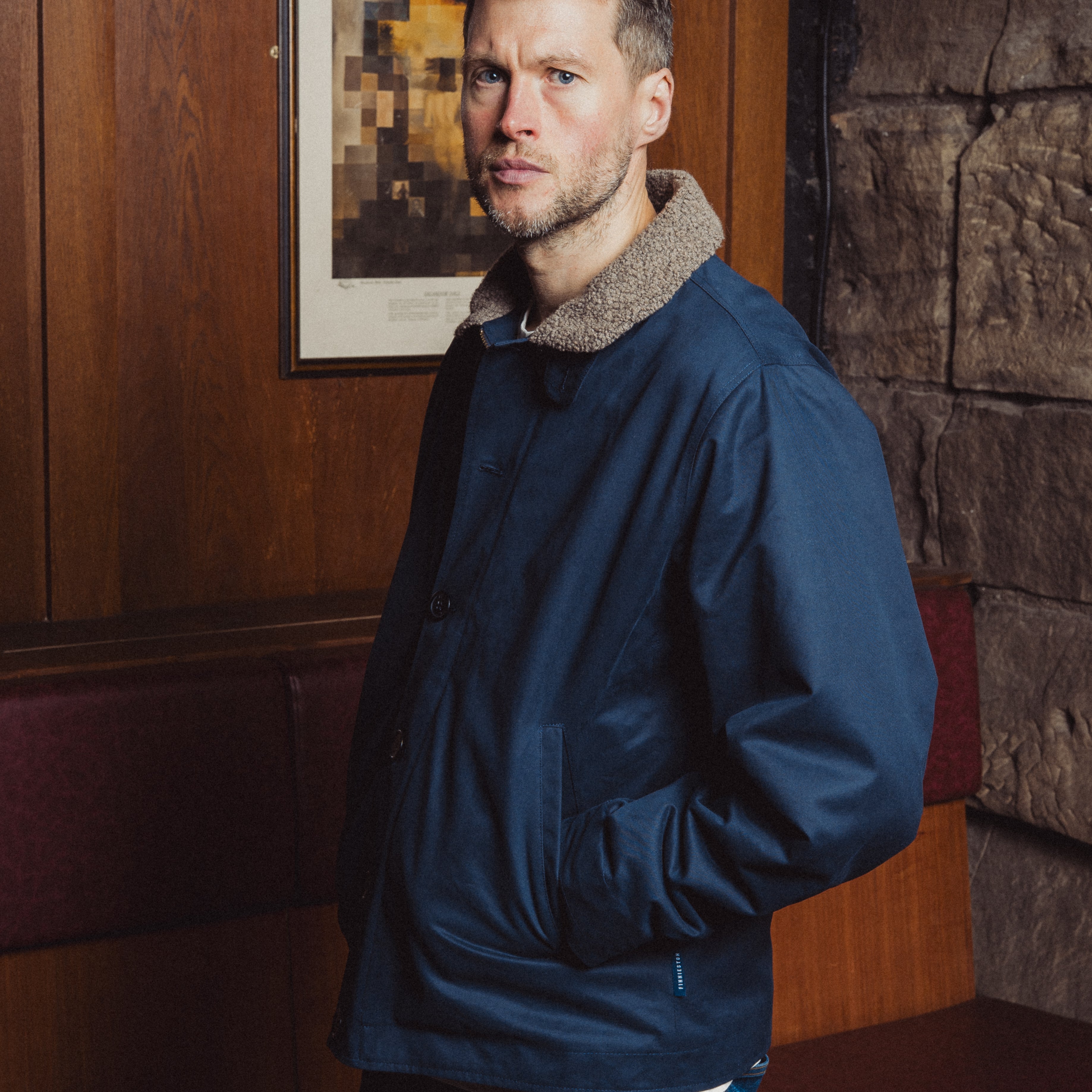 Man wearing a blue jacket in a room with wooden paneling and a painting on the wall.