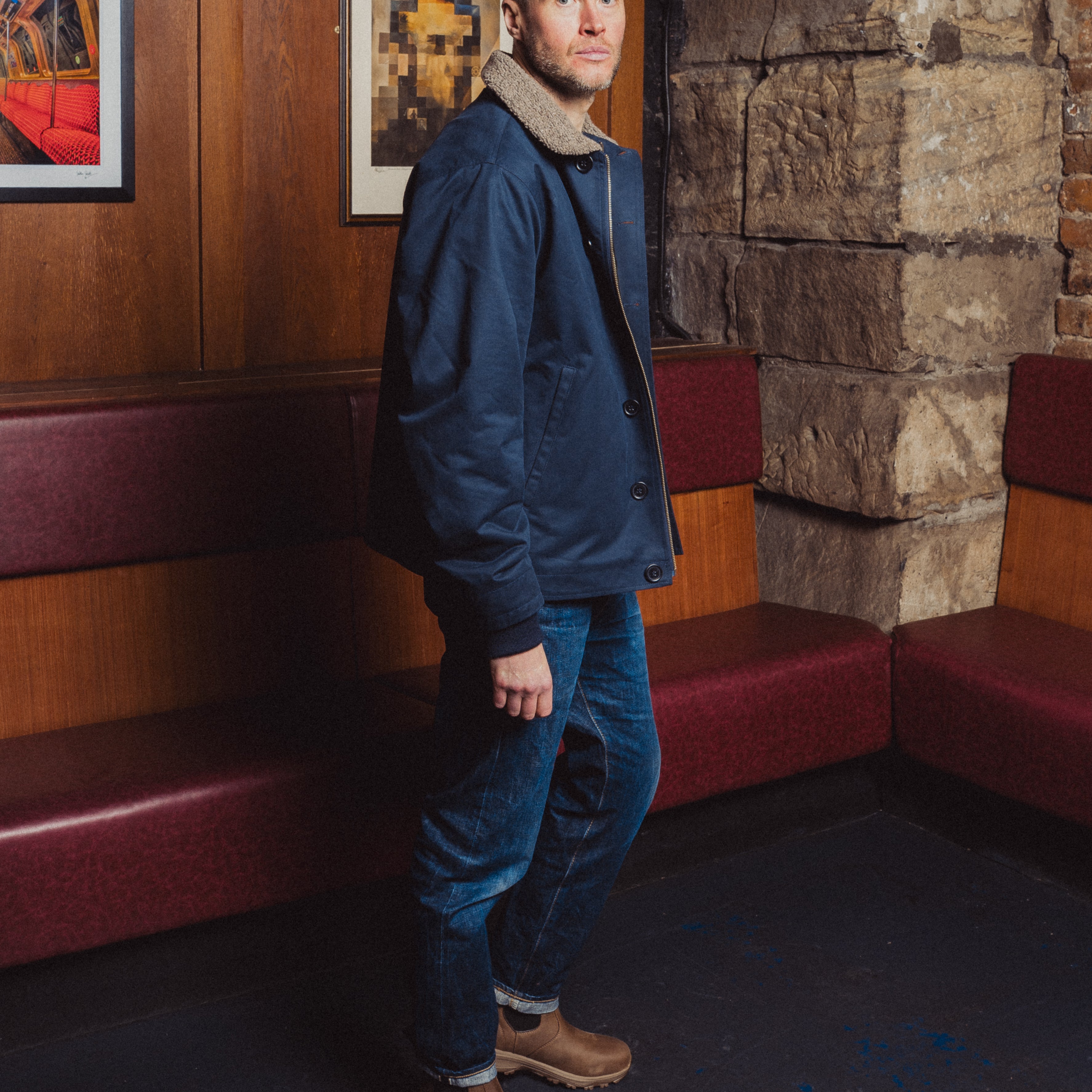 Man in a blue jacket standing in a room with wooden paneling and framed pictures.