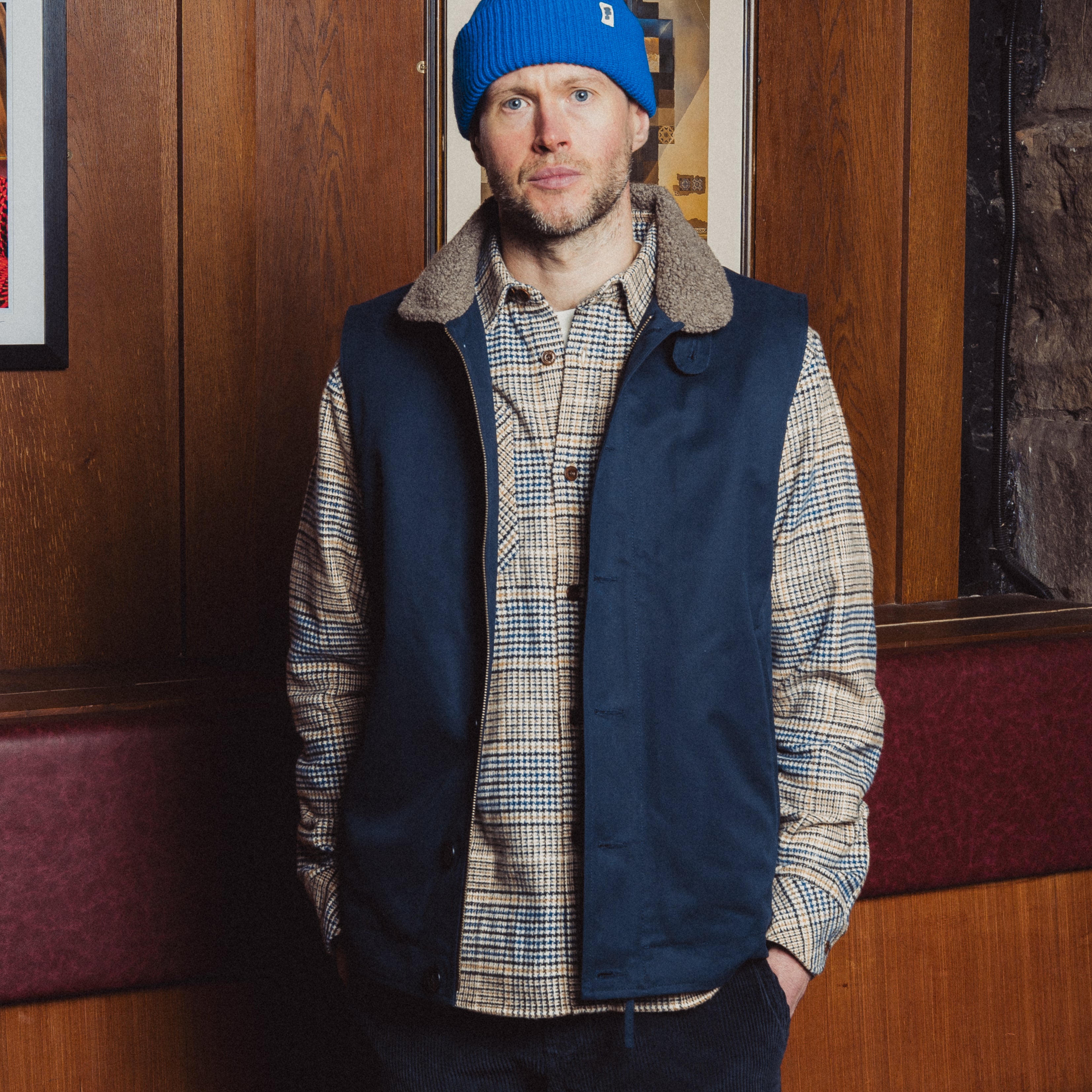Man wearing a blue beanie and navy vest over a plaid shirt in an indoor setting.