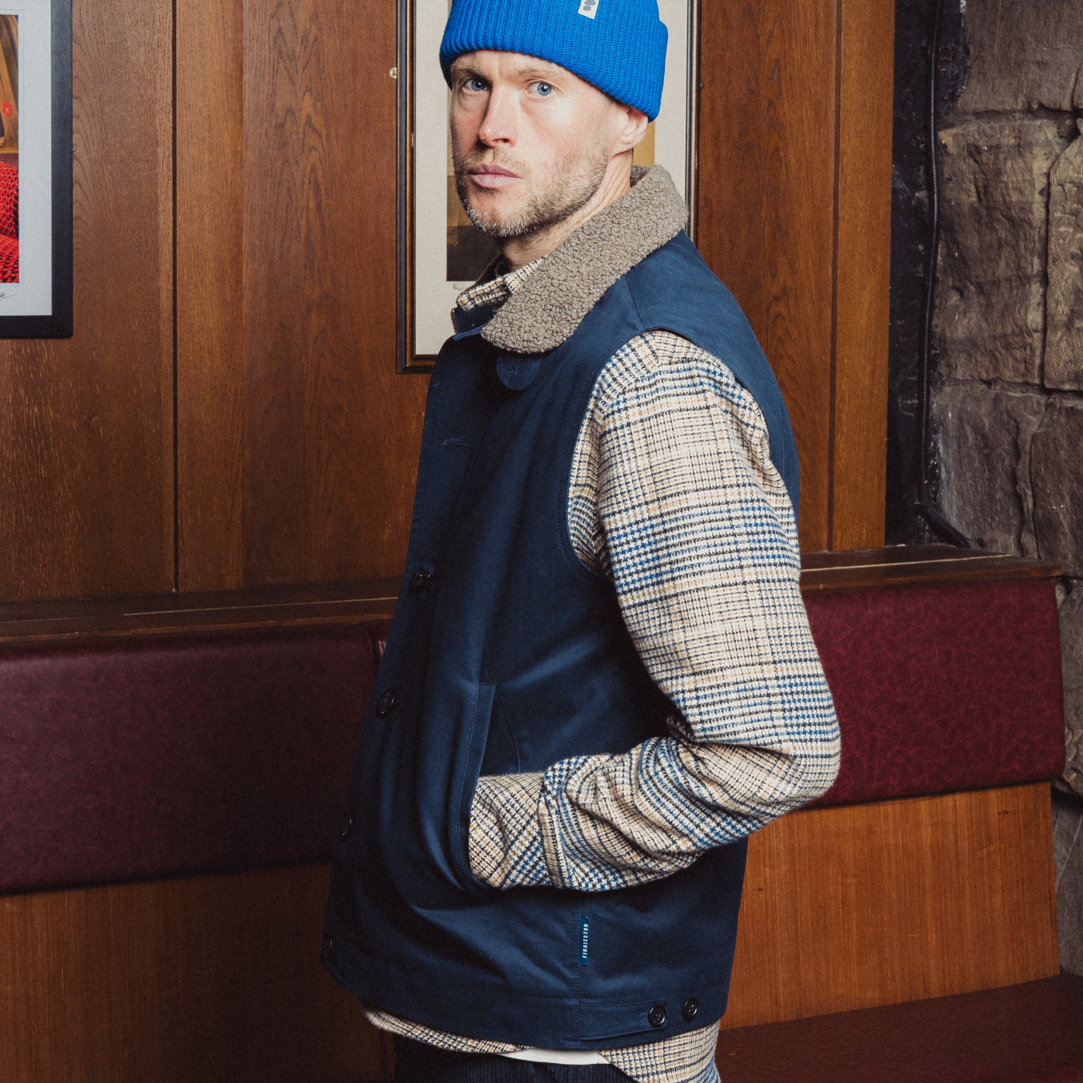 Man wearing a blue beanie and navy vest in a room with wooden paneling and framed pictures.