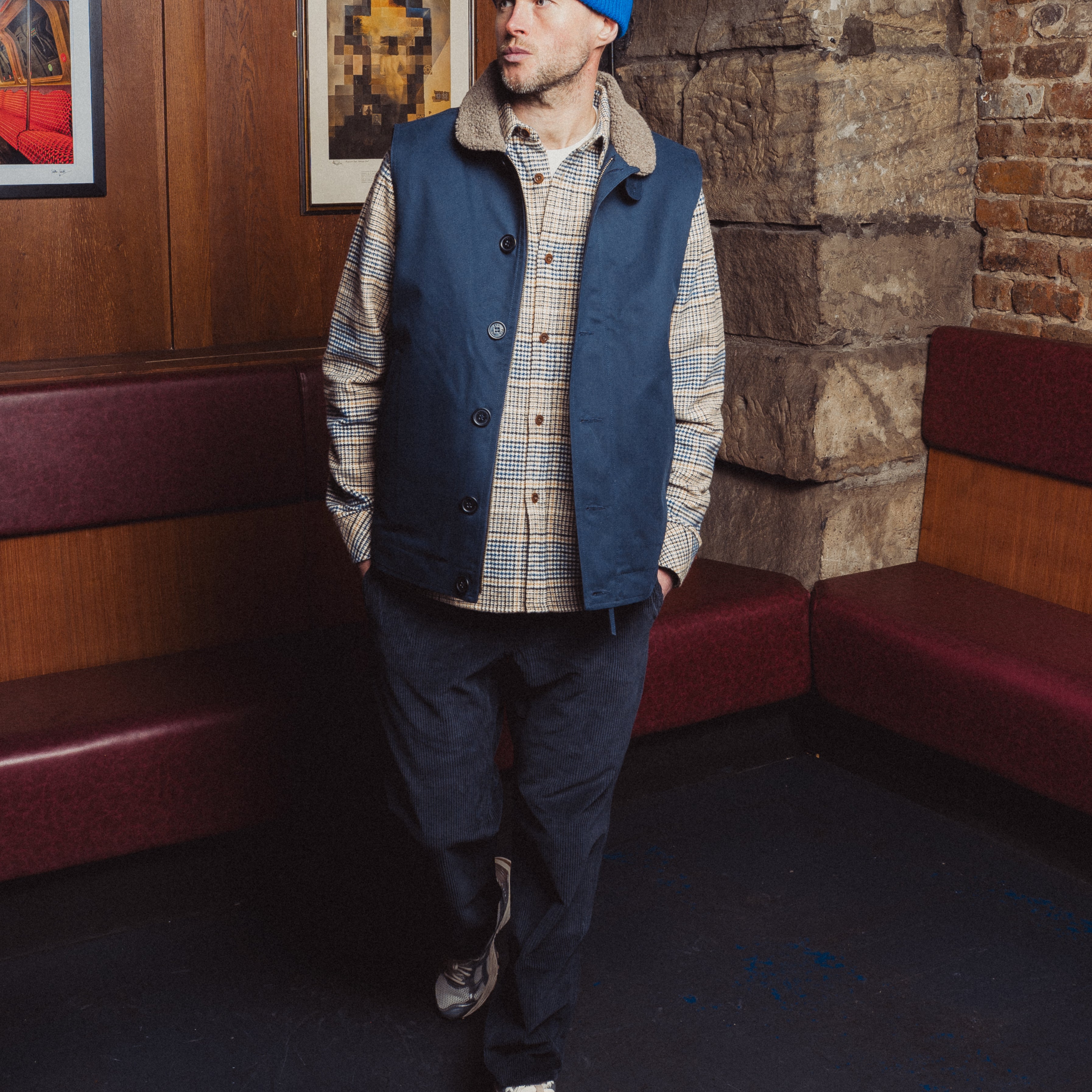 Man wearing a blue vest and plaid shirt standing in a room with brick walls and framed pictures.