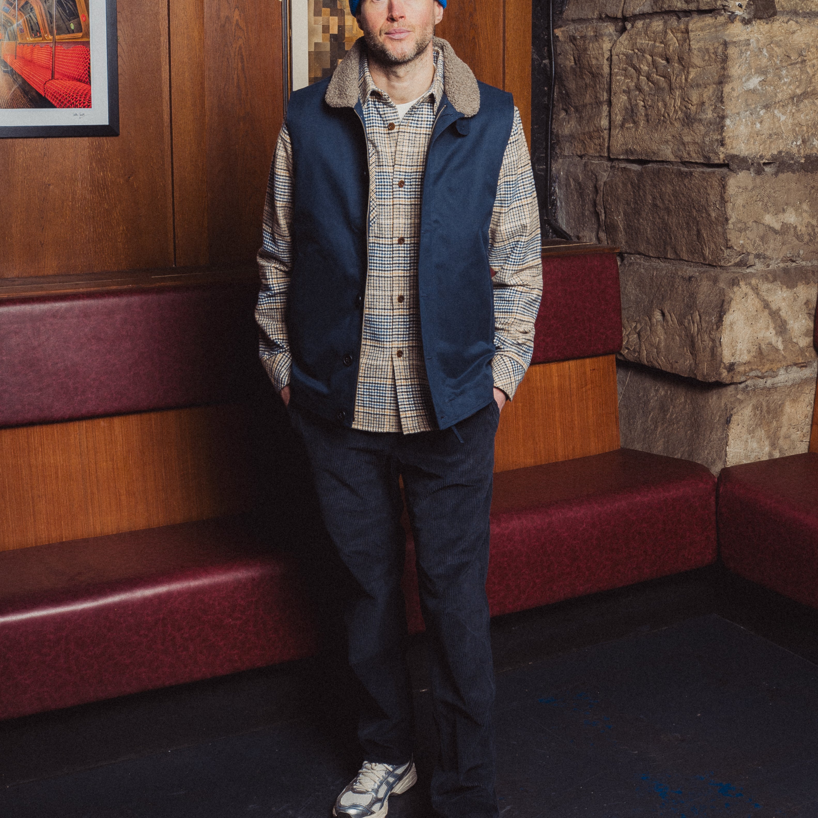 Man wearing a blue beanie and vest standing in a room with wooden walls and red benches.