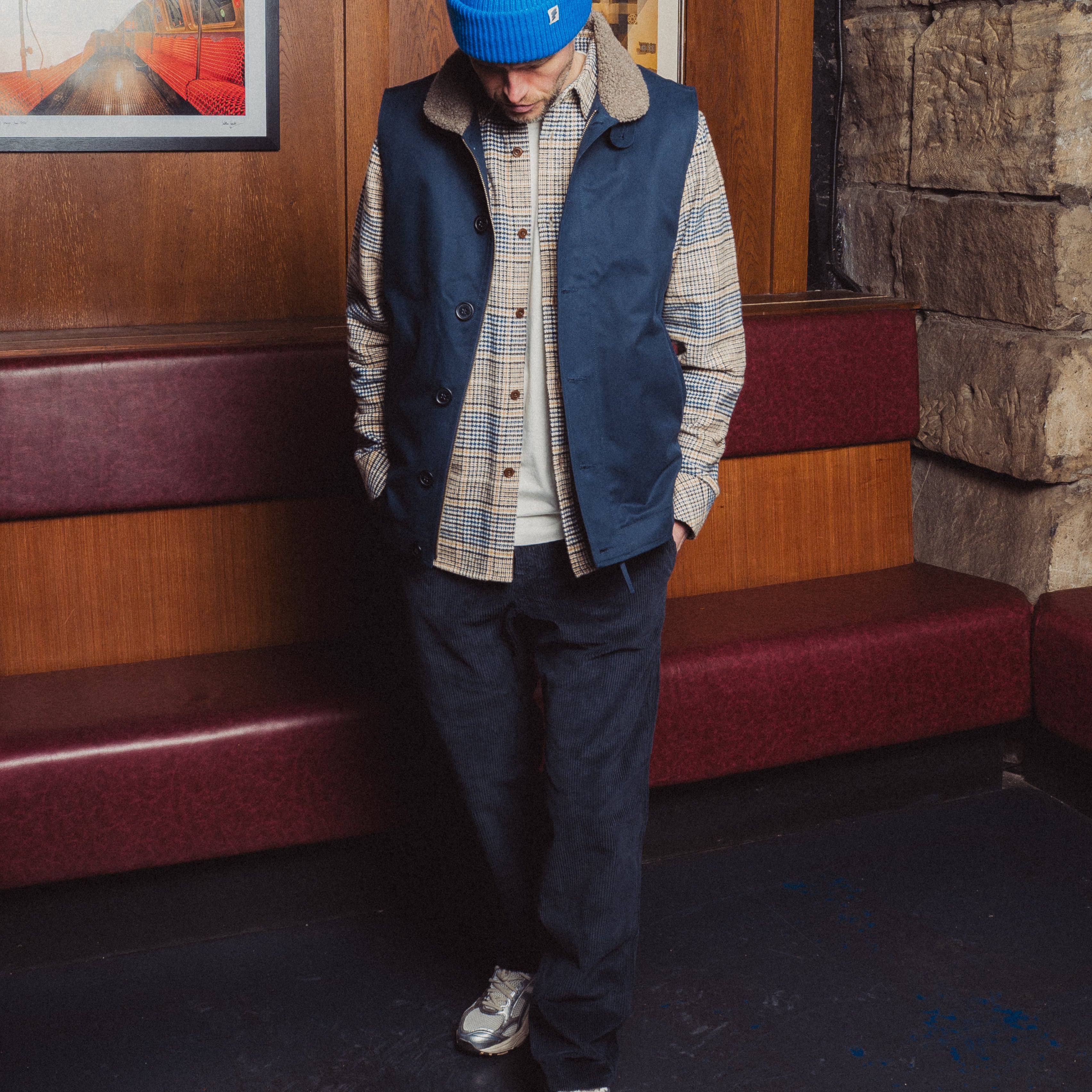 Person wearing a blue vest, plaid shirt, and dark pants standing in a room with wooden walls and a stone wall.