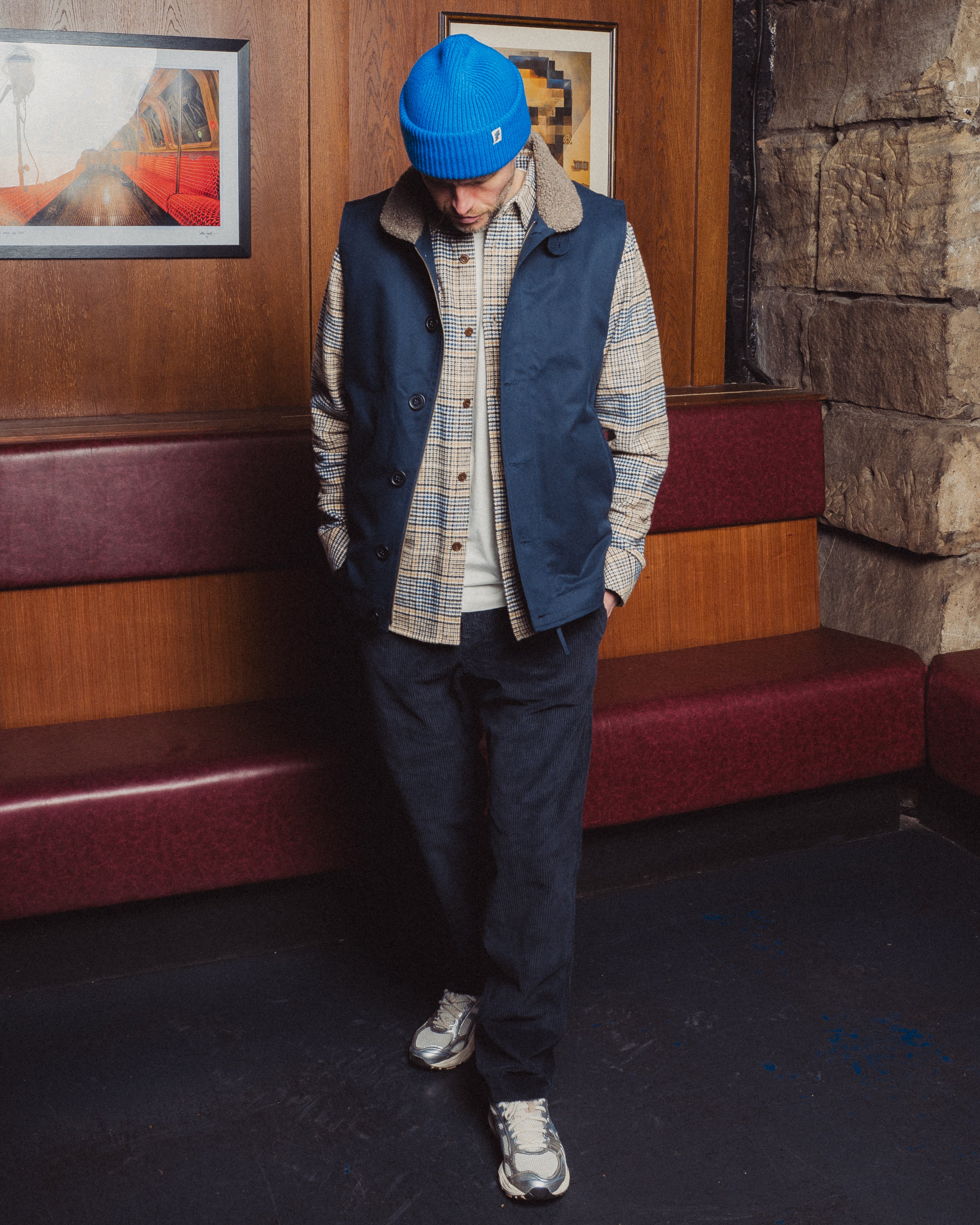 Person wearing a blue vest, plaid shirt, and dark pants standing in a room with wooden walls and a stone wall.