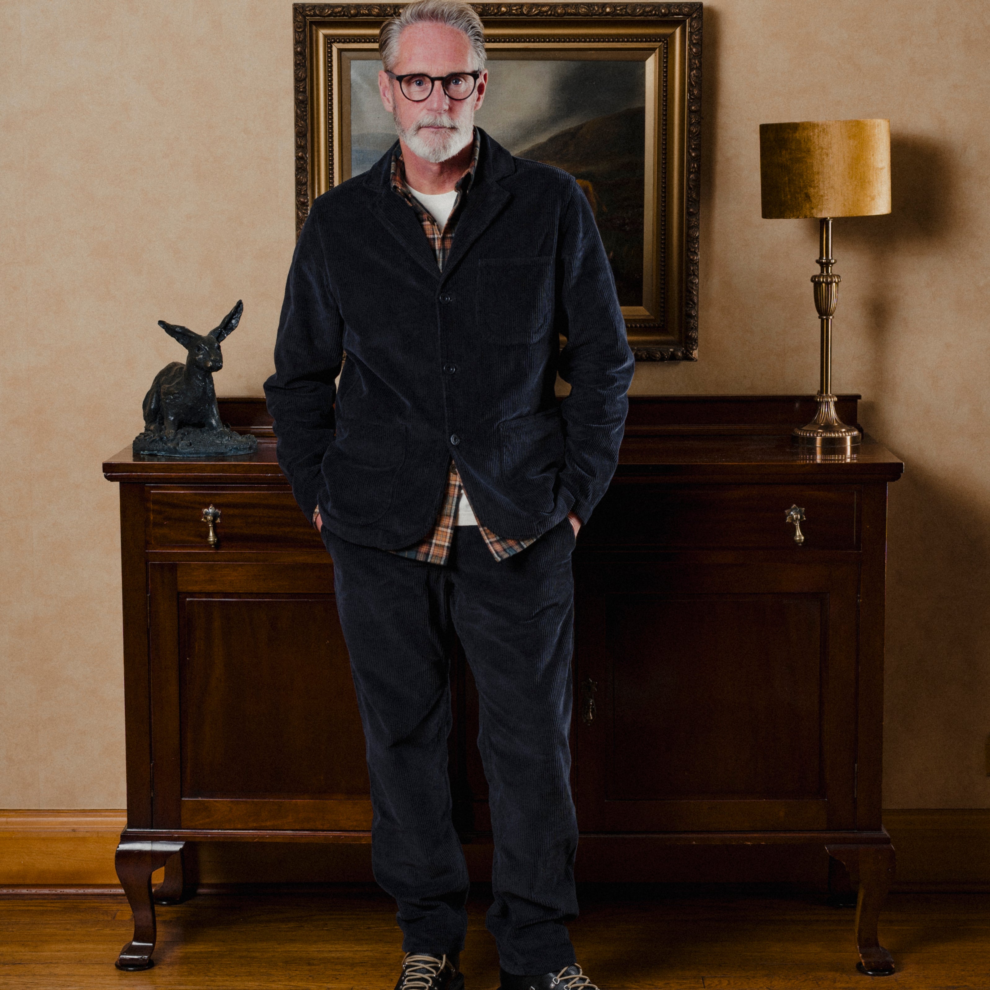 Man wearing navy corduroy suit standing in a room with a painting and lamp on a wall.