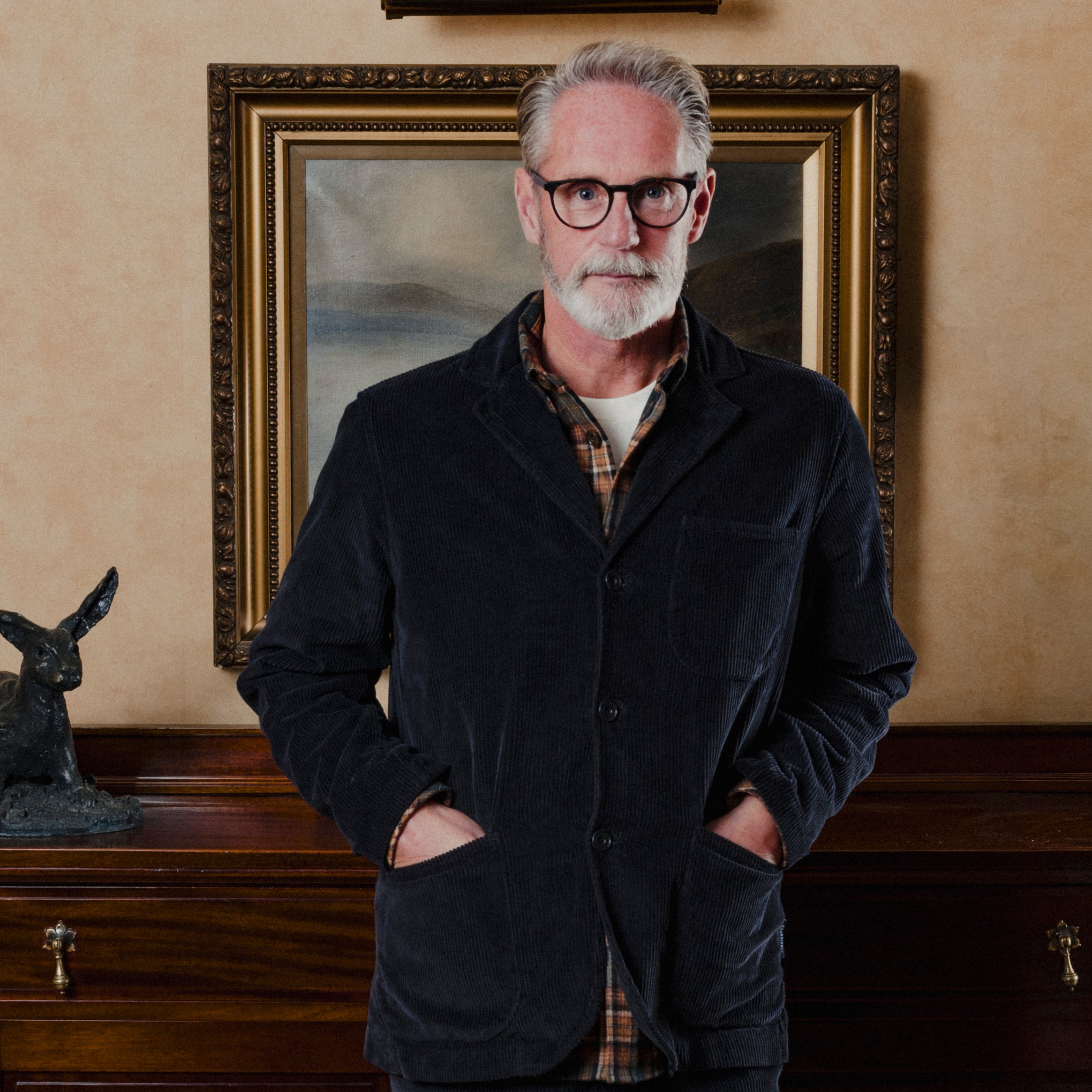 Man wearing a navy corduroy suit jacket and glasses standing in front of a framed painting on a beige wall.