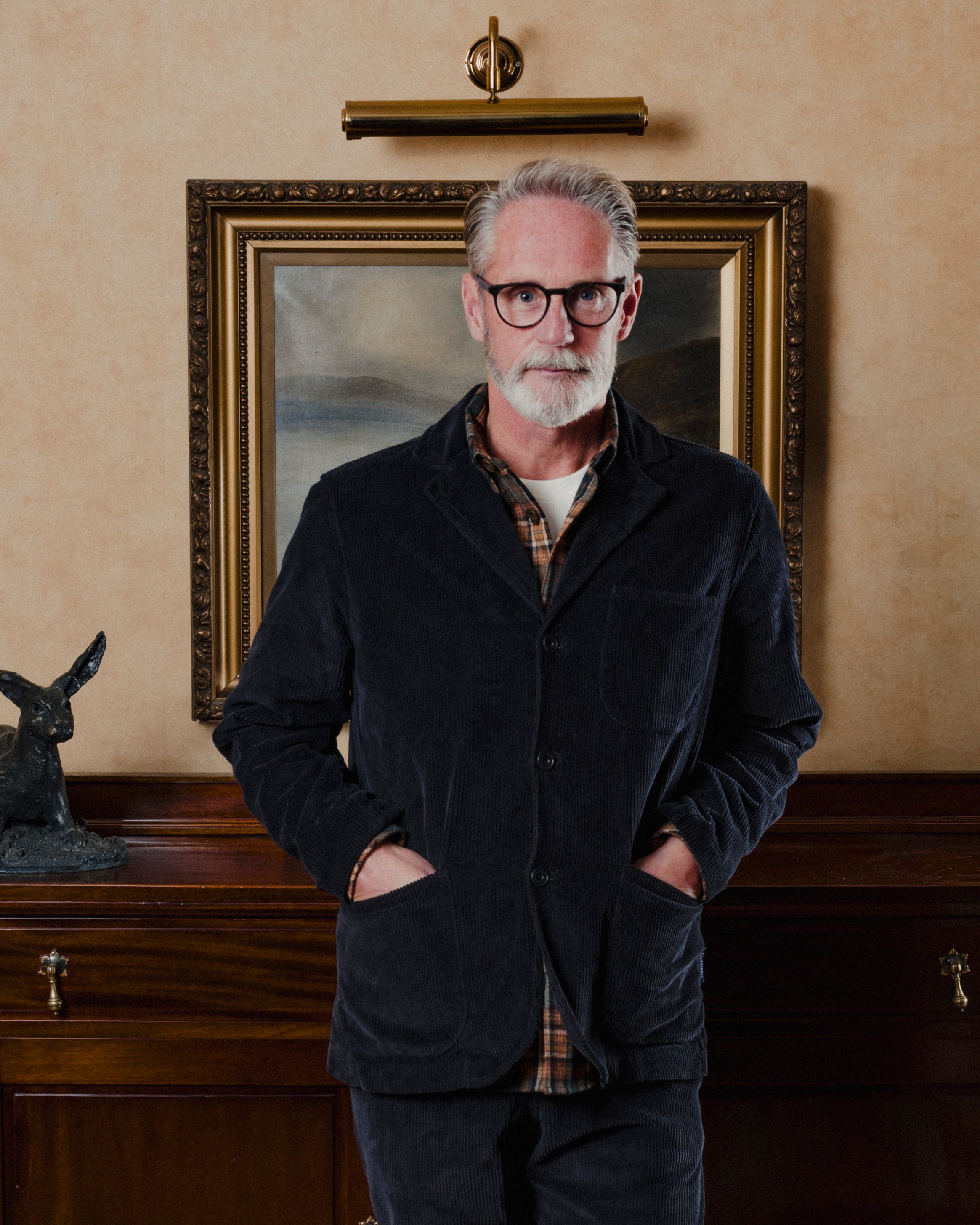 Man wearing a navy corduroy suit jacket and glasses standing in front of a framed painting on a beige wall.