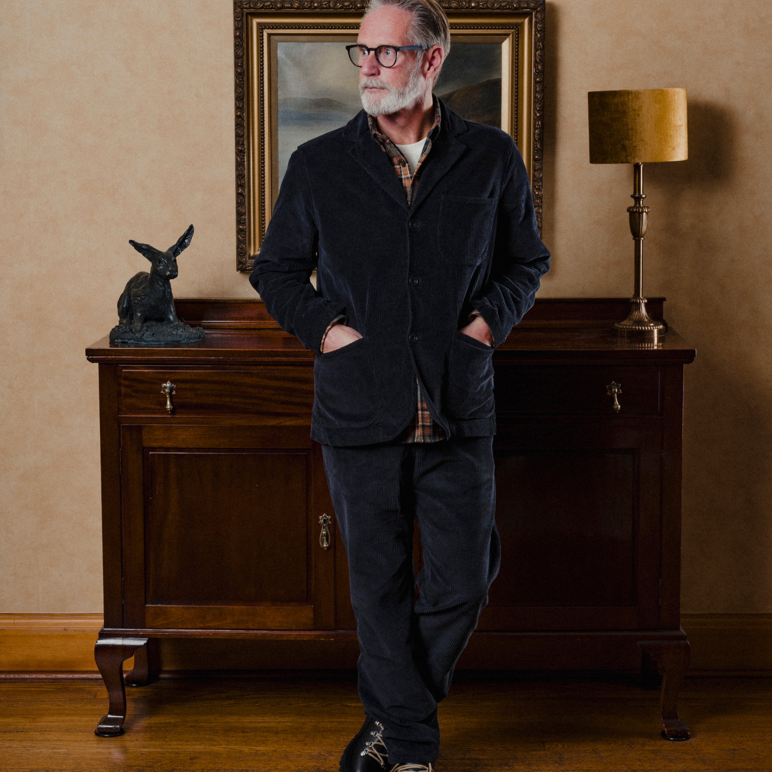 Man in a dark suit standing in a room with a wooden cabinet, lamp, and painting.