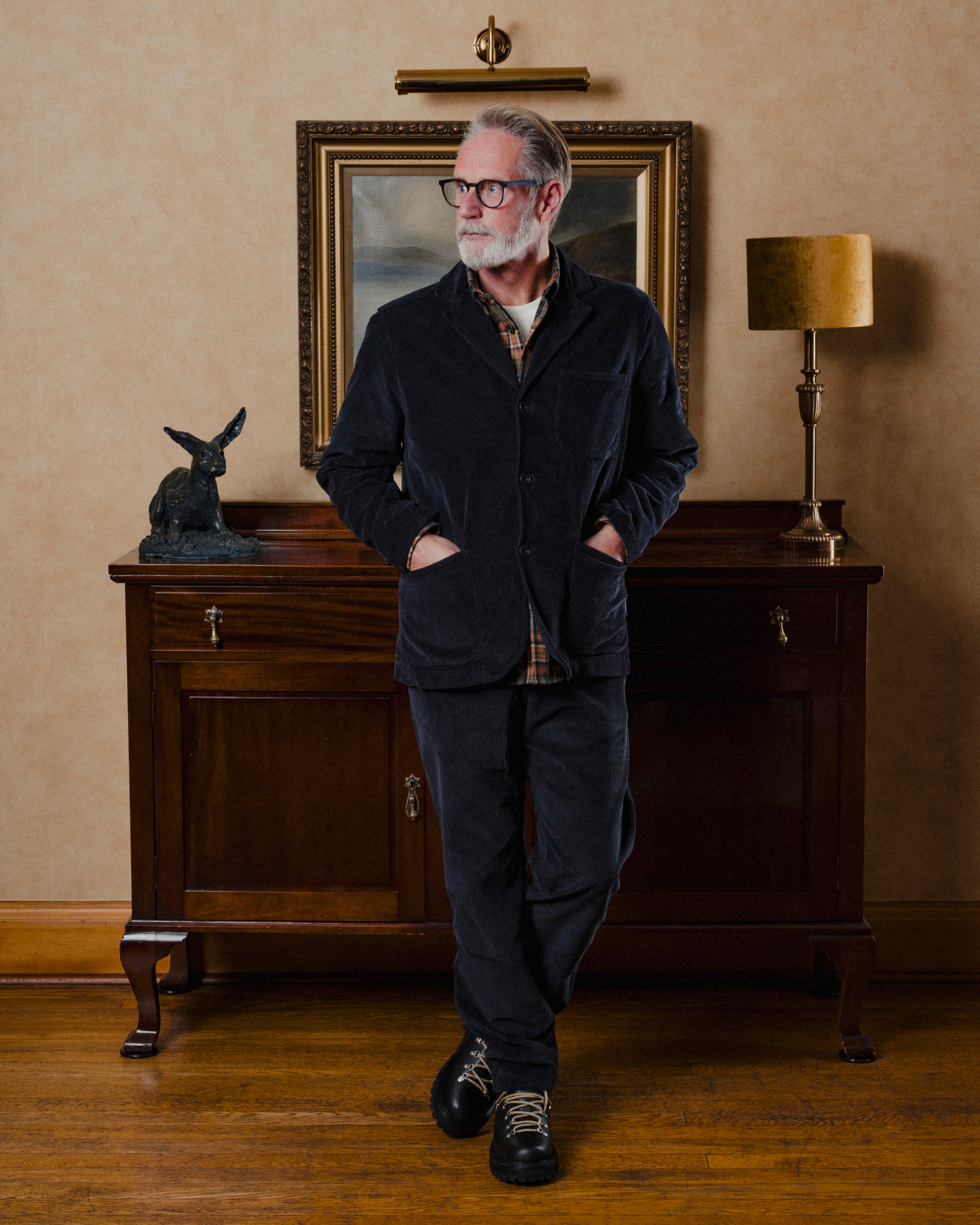 Man in a dark suit standing in a room with a wooden cabinet, lamp, and painting.
