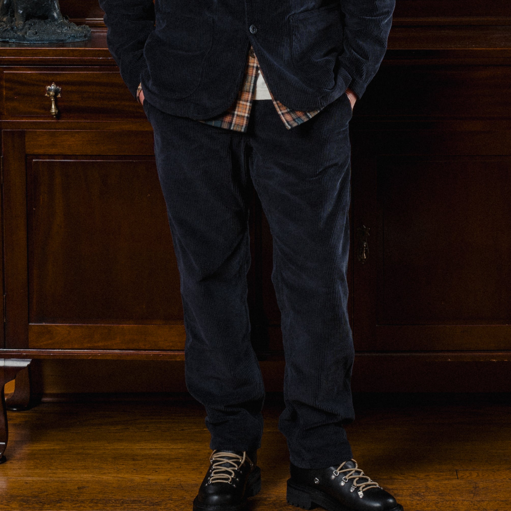 Person wearing a dark jacket, plaid shirt, and dark pants standing in a room with wooden furniture.