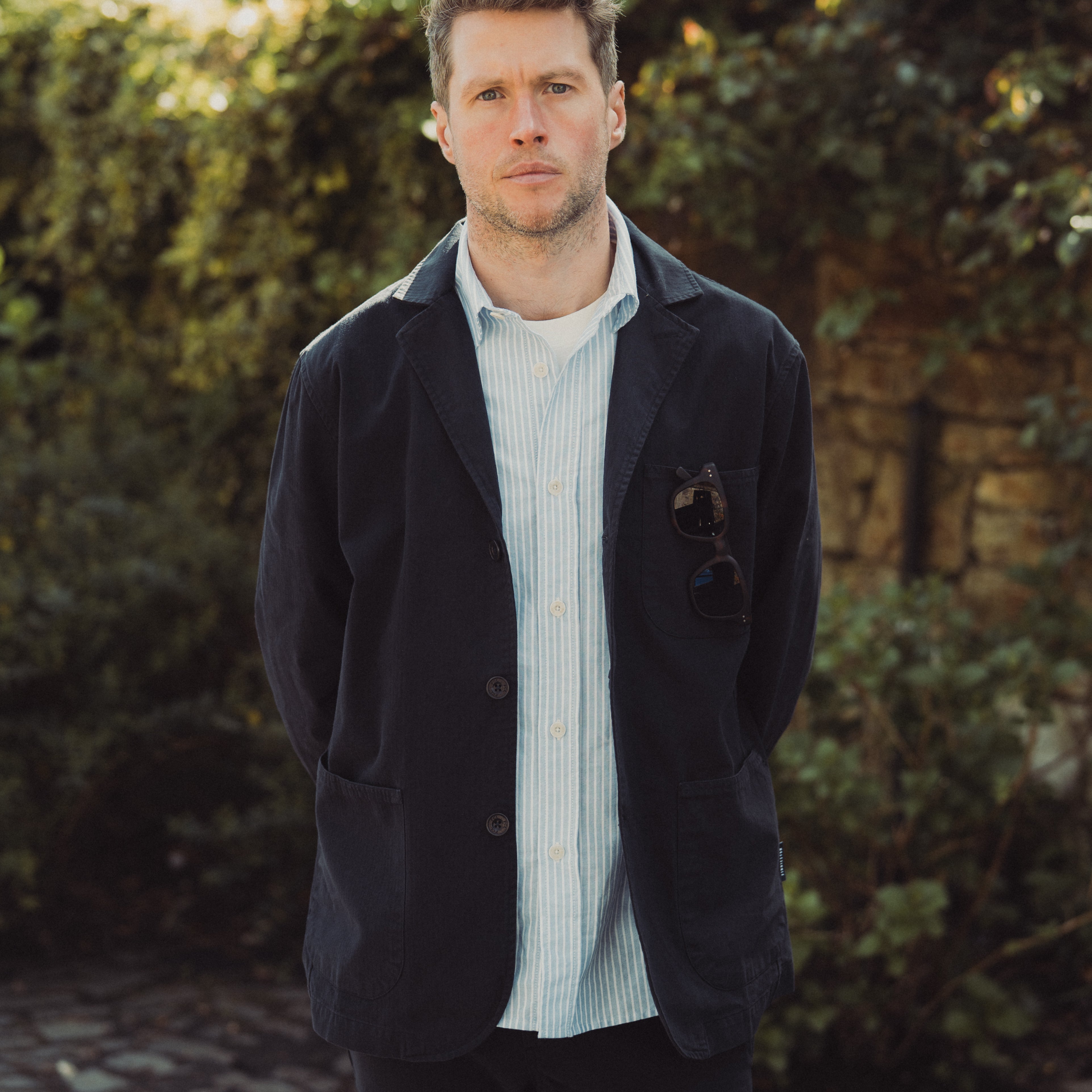 Man wearing a dark jacket over a light shirt standing outdoors with greenery in the background