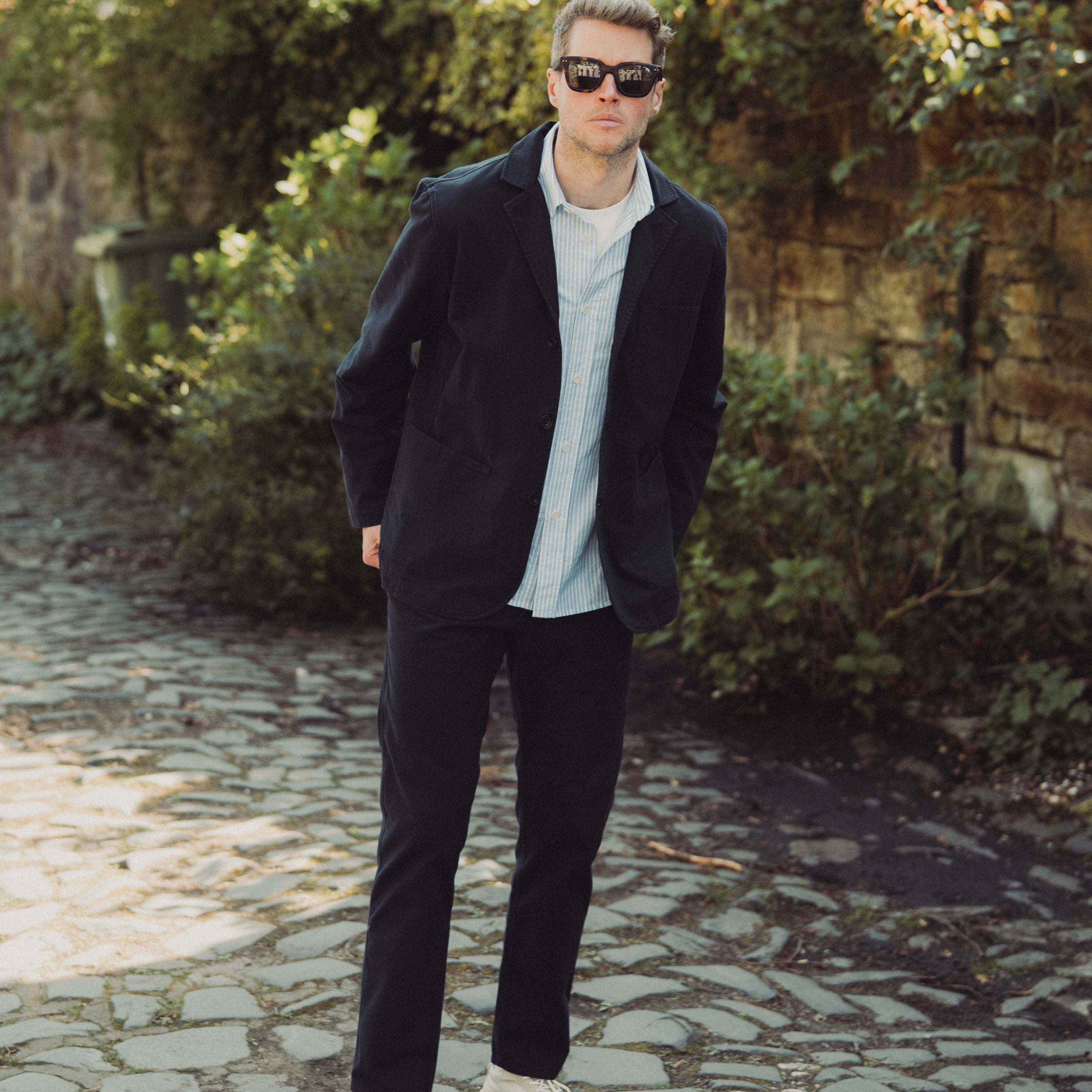 Man in dark coat and sunglasses standing on a stone path with greenery in the background