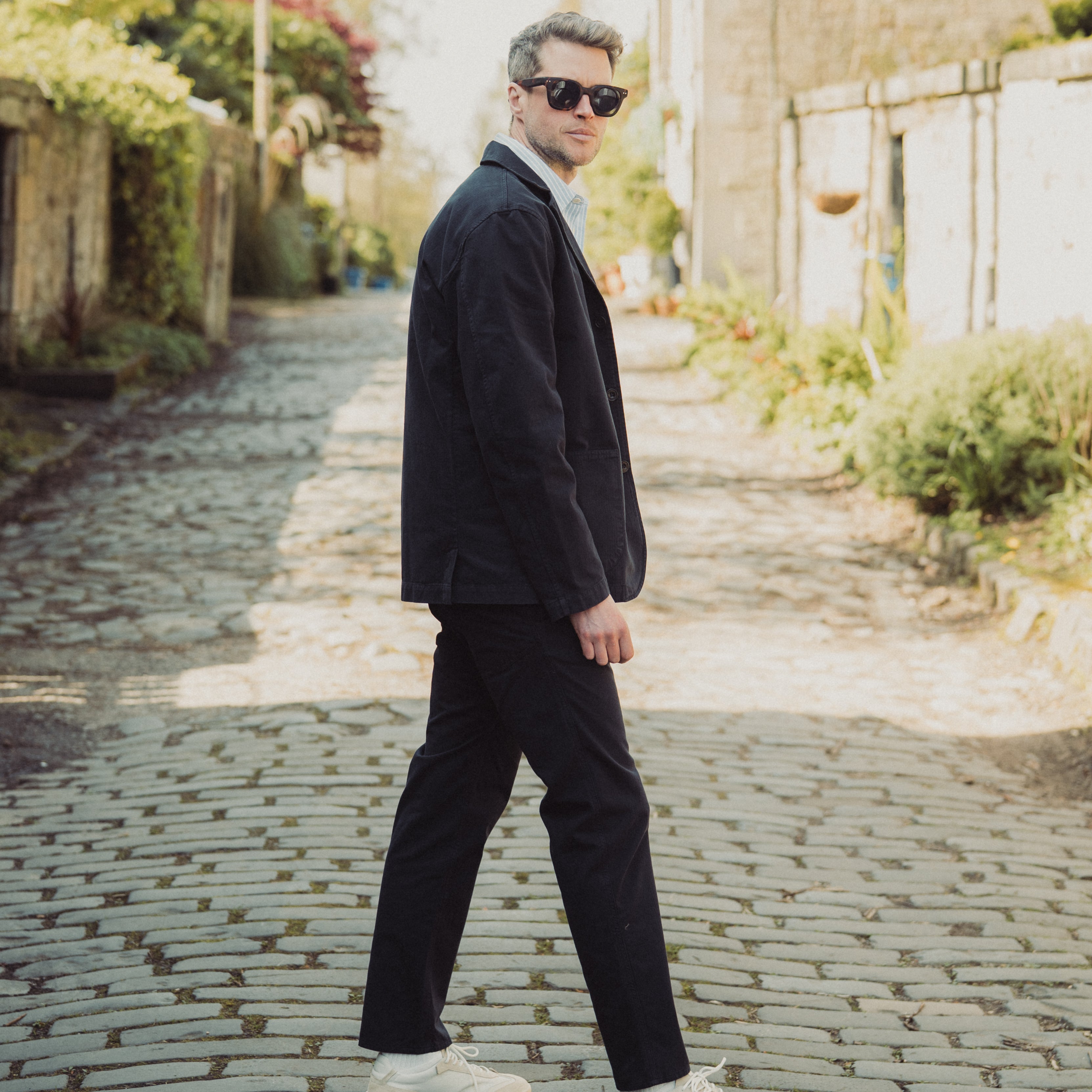 Man in a suit walking on a cobblestone street with greenery and a building in the background