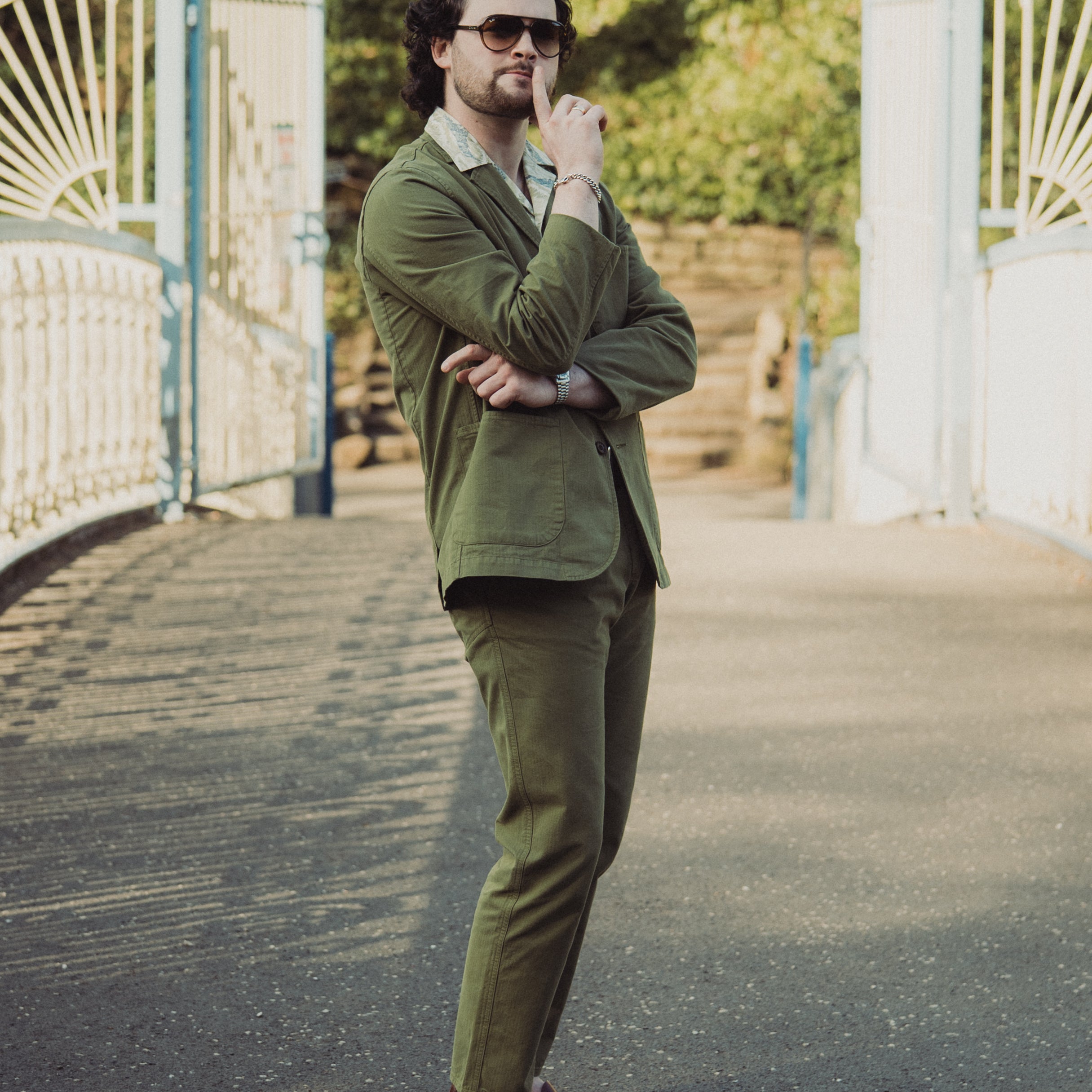 Man in a green suit standing on a pathway with white gates and greenery in the background