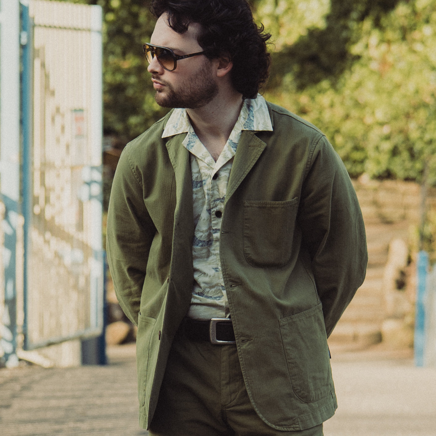 Man wearing a green jacket and sunglasses outdoors with greenery in the background