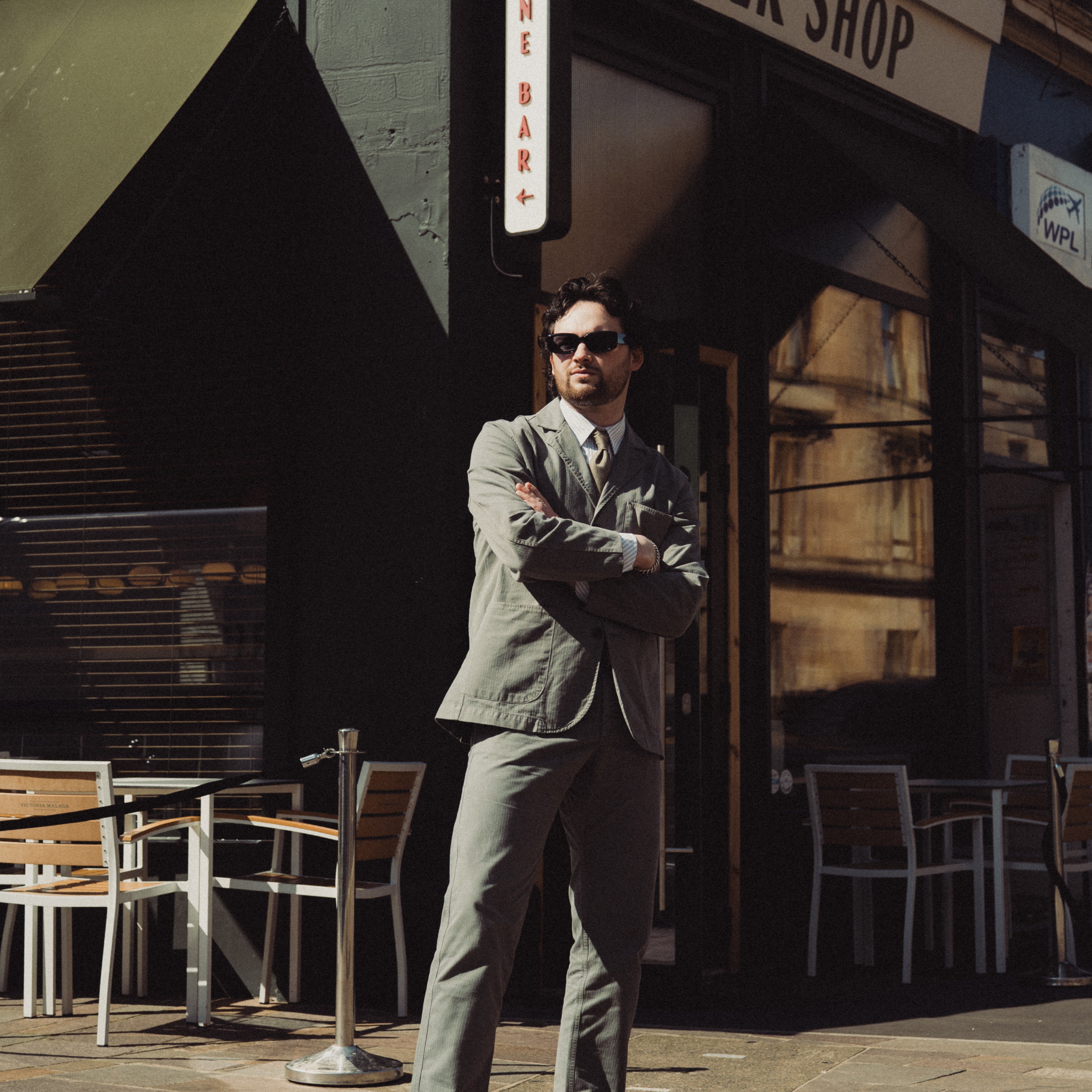 Man in a suit standing outside a corner shop with tables and chairs.
