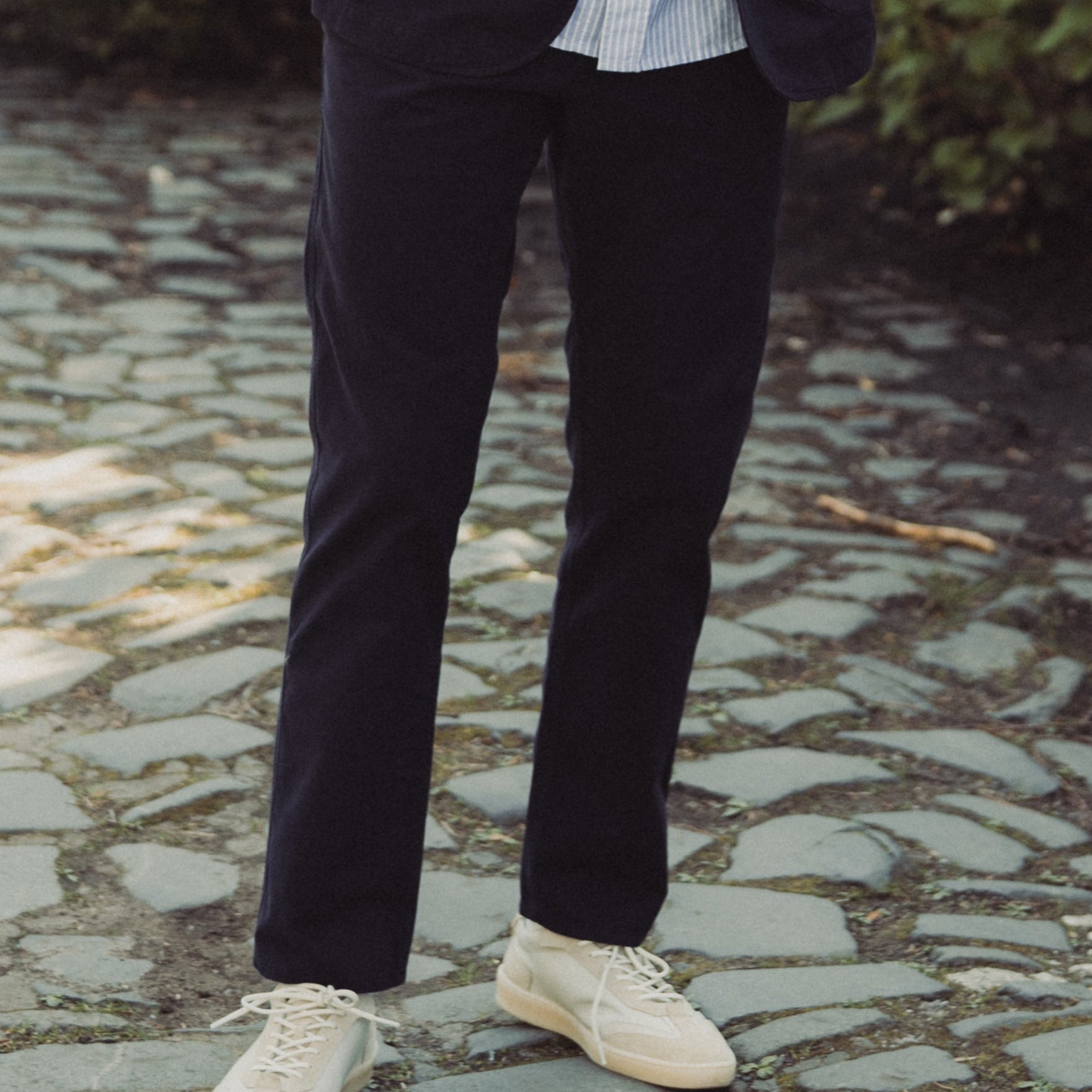 Man in dark coat and sunglasses standing on a stone path with greenery in the background