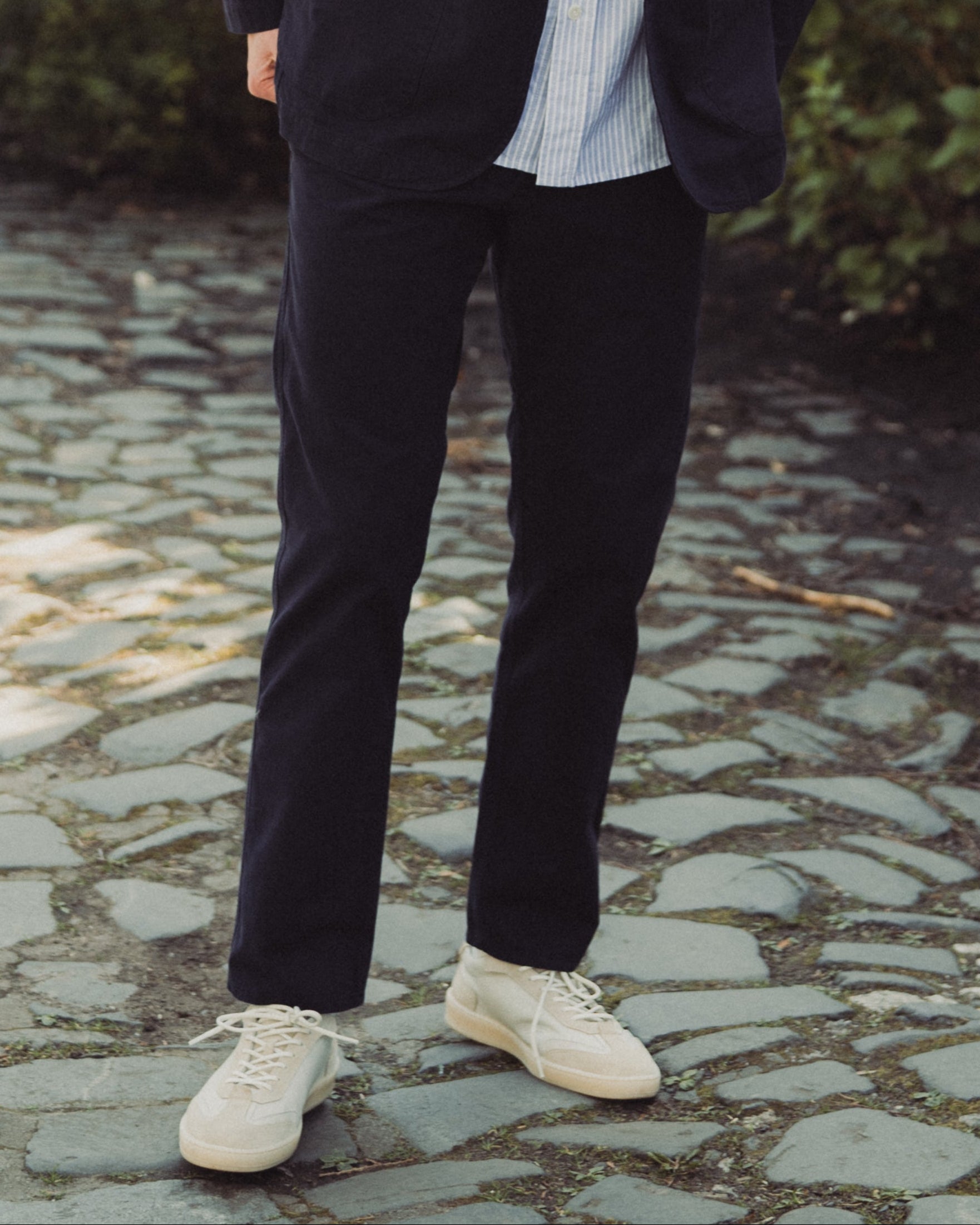Man in dark coat and sunglasses standing on a stone path with greenery in the background