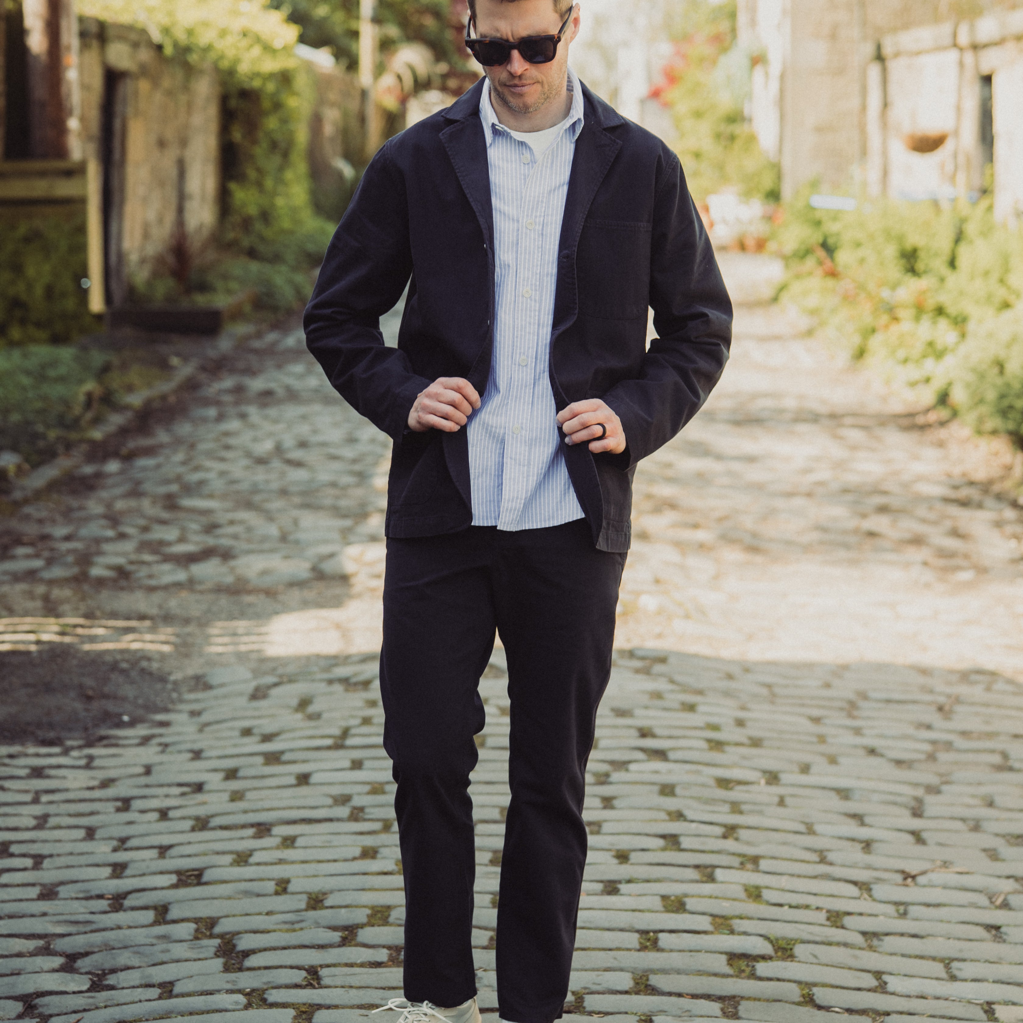 Man wearing a dark jacket, light shirt, and white sneakers on a cobblestone street.