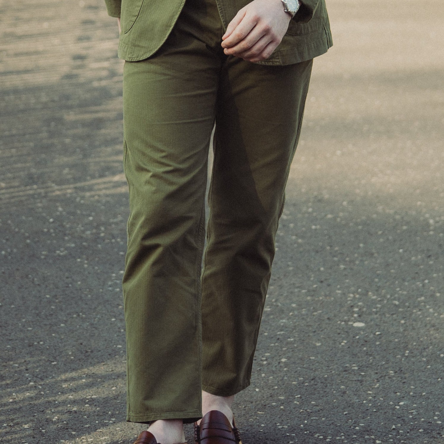 Man in a green jacket and pants standing on a paved path with a white gate in the background.