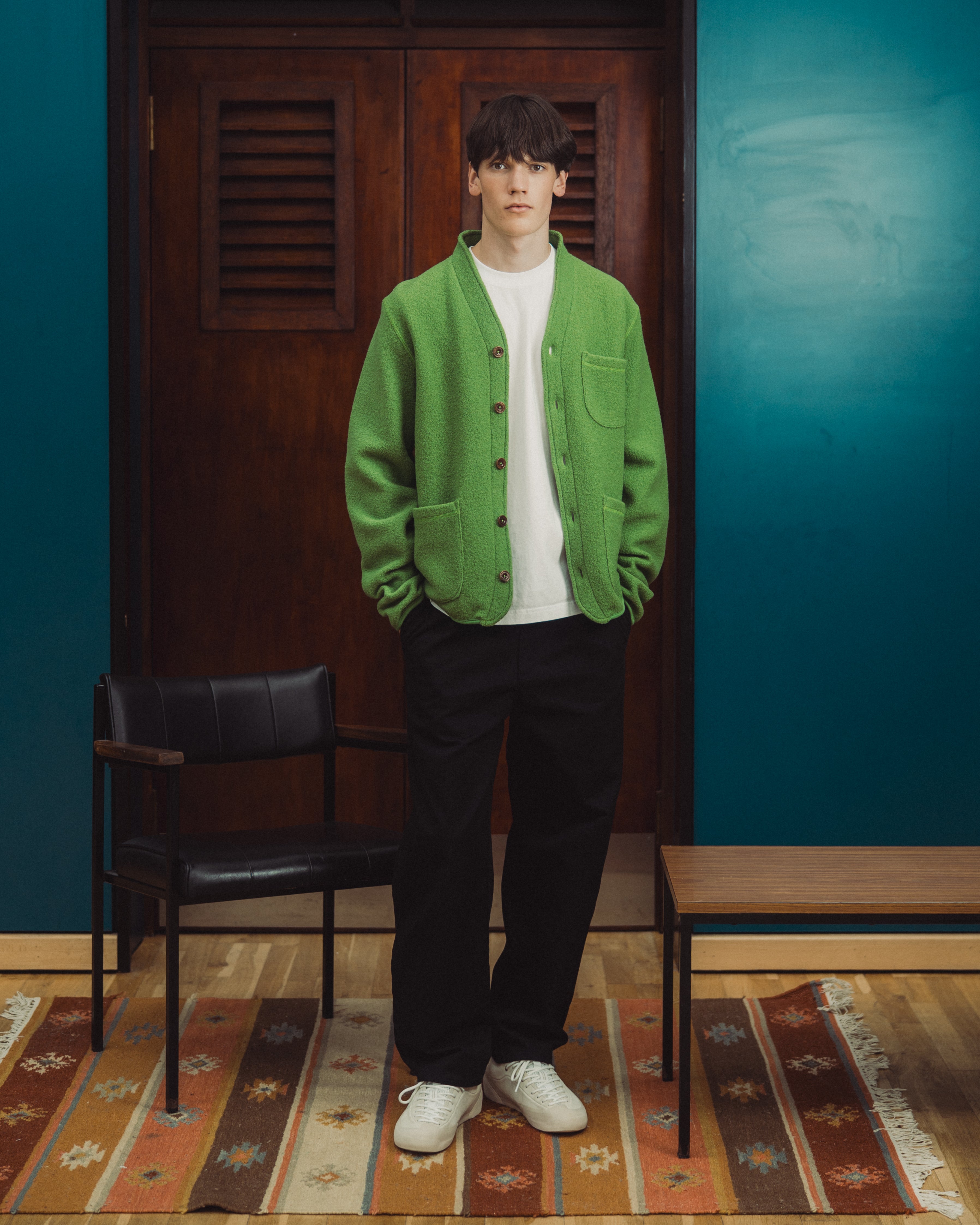 A man stands in a room wearing a bright green knit cardigan over a white turtleneck, black trousers, and white sneakers. The background features a dark wood door, teal-blue walls, a black chair, and a patterned rug.