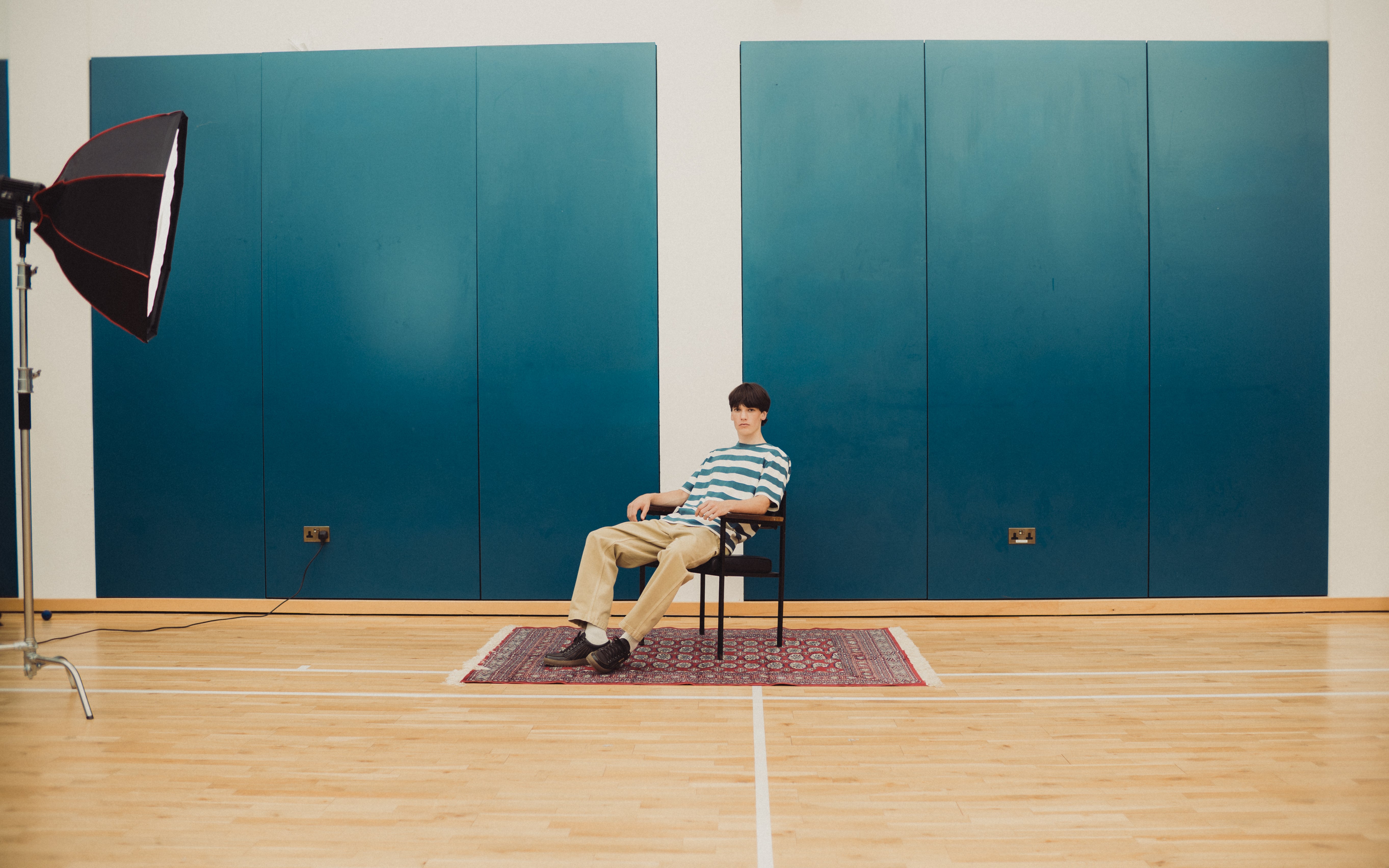 A man sits on a chair on a patterned rug in a studio with blue wall panels and a wooden floor. A softbox light stand is visible on the left.