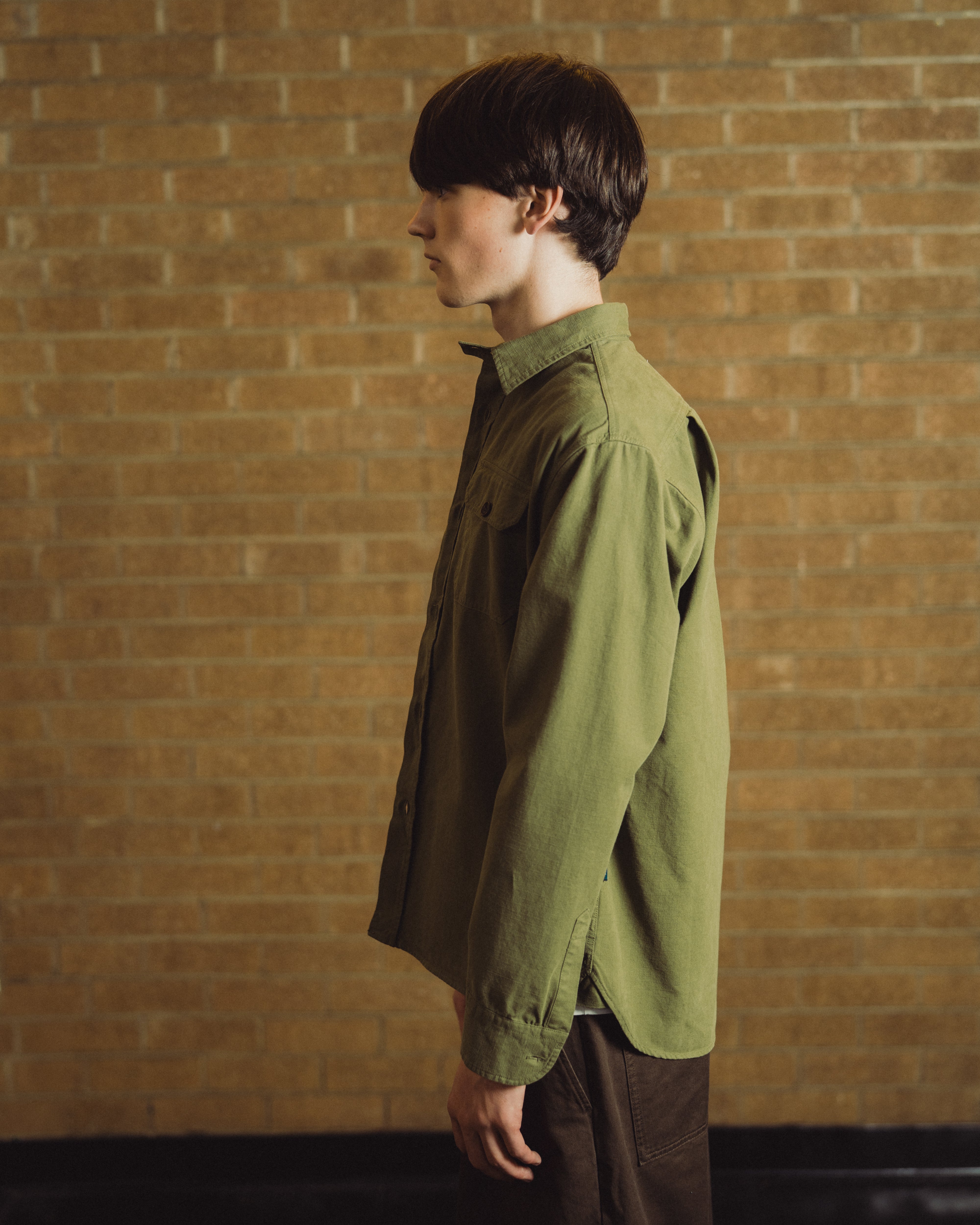 A man in profile wearing an olive green button-up shirt and dark brown trousers, standing against a warm tan brick wall.