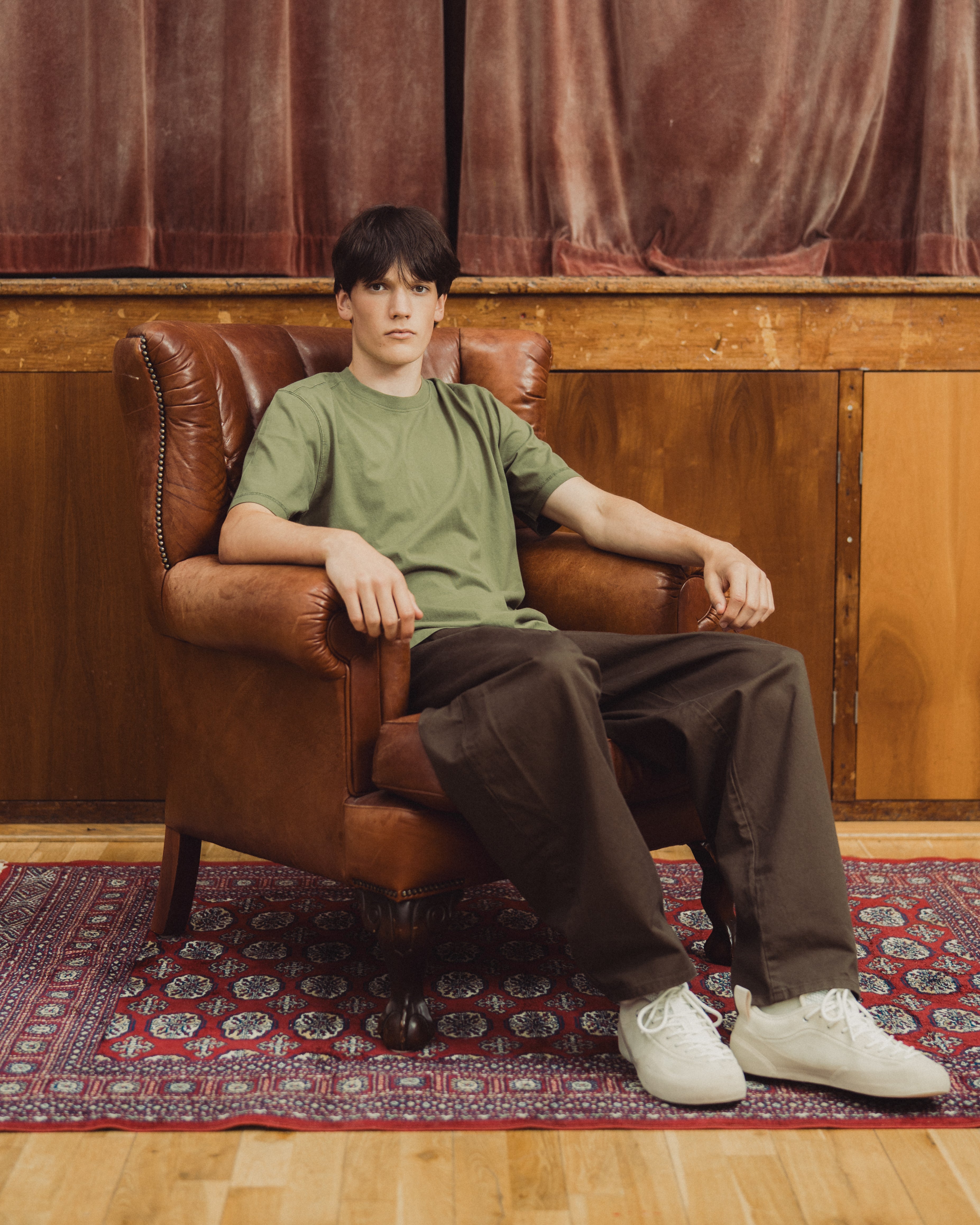 A young man sits in a brown leather armchair wearing an olive green t-shirt, dark brown pants, and white sneakers, set against a background of wood paneling and a patterned rug.